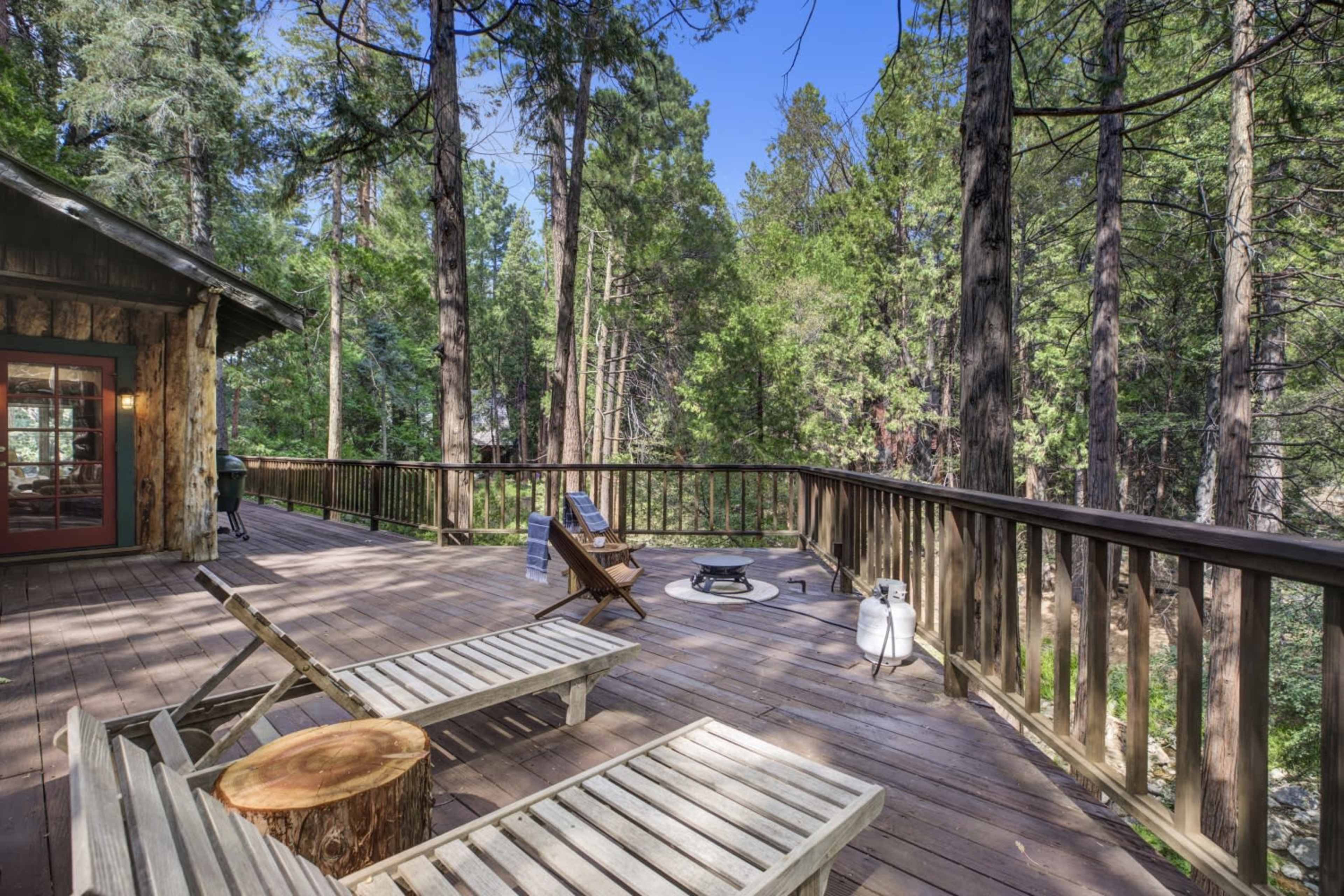 A wooden deck is surrounded by tall trees, featuring lounge chairs and a fire pit.