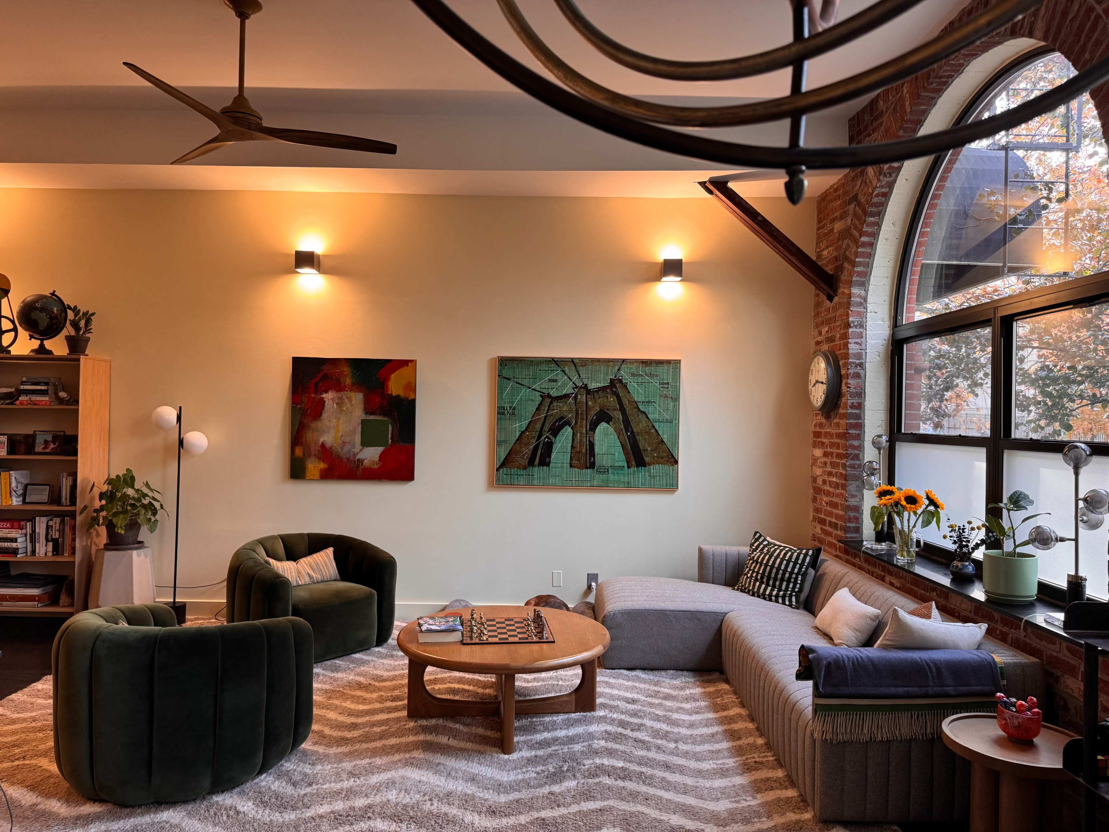 A warmly lit living room features a mix of seating, a round coffee table, and large windows adorned with sunflowers, with artwork hanging on the walls.