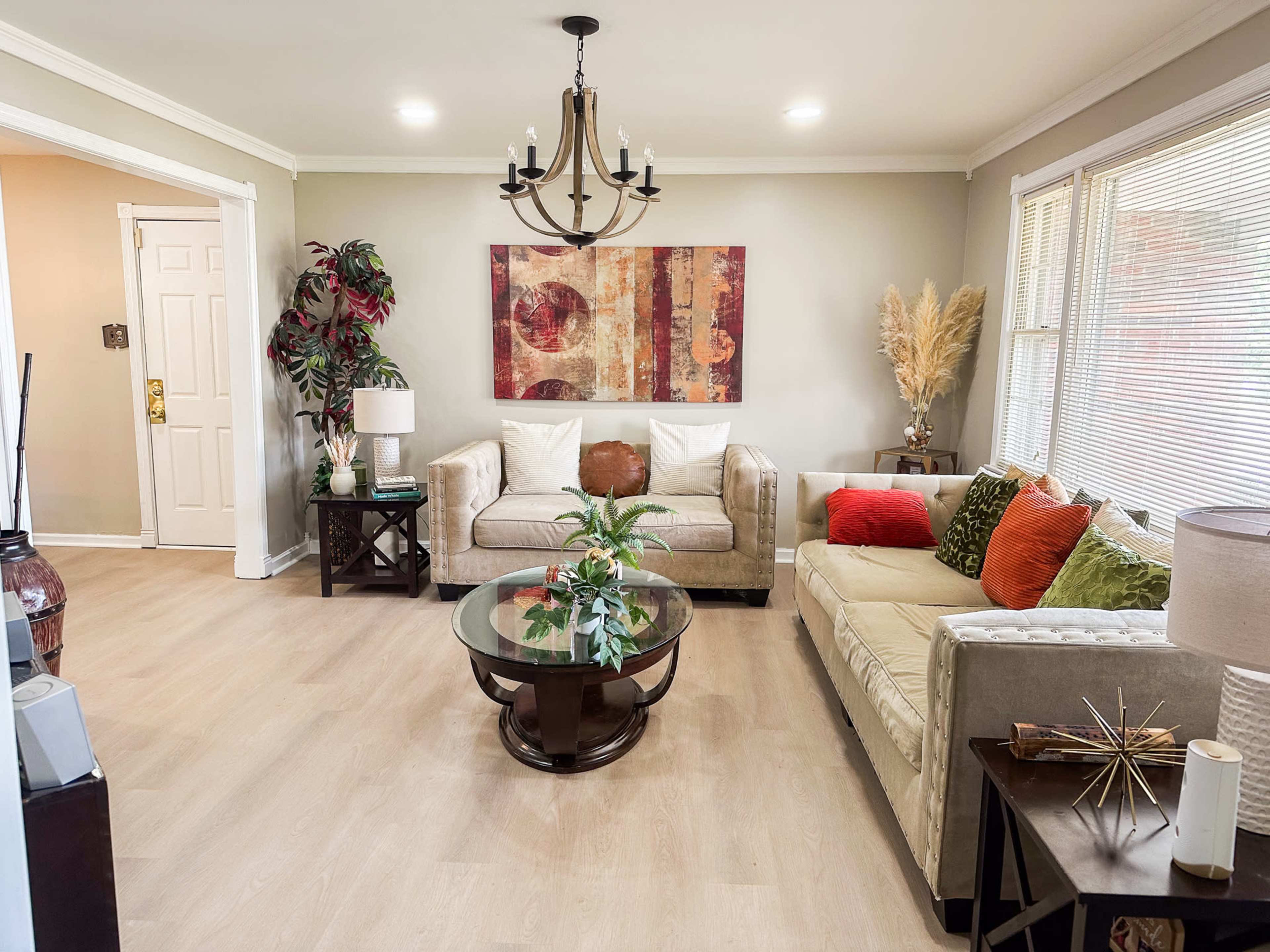 The image shows a living room with two couches, a coffee table, decorative plants, and a wall art piece, illuminated by overhead lighting.