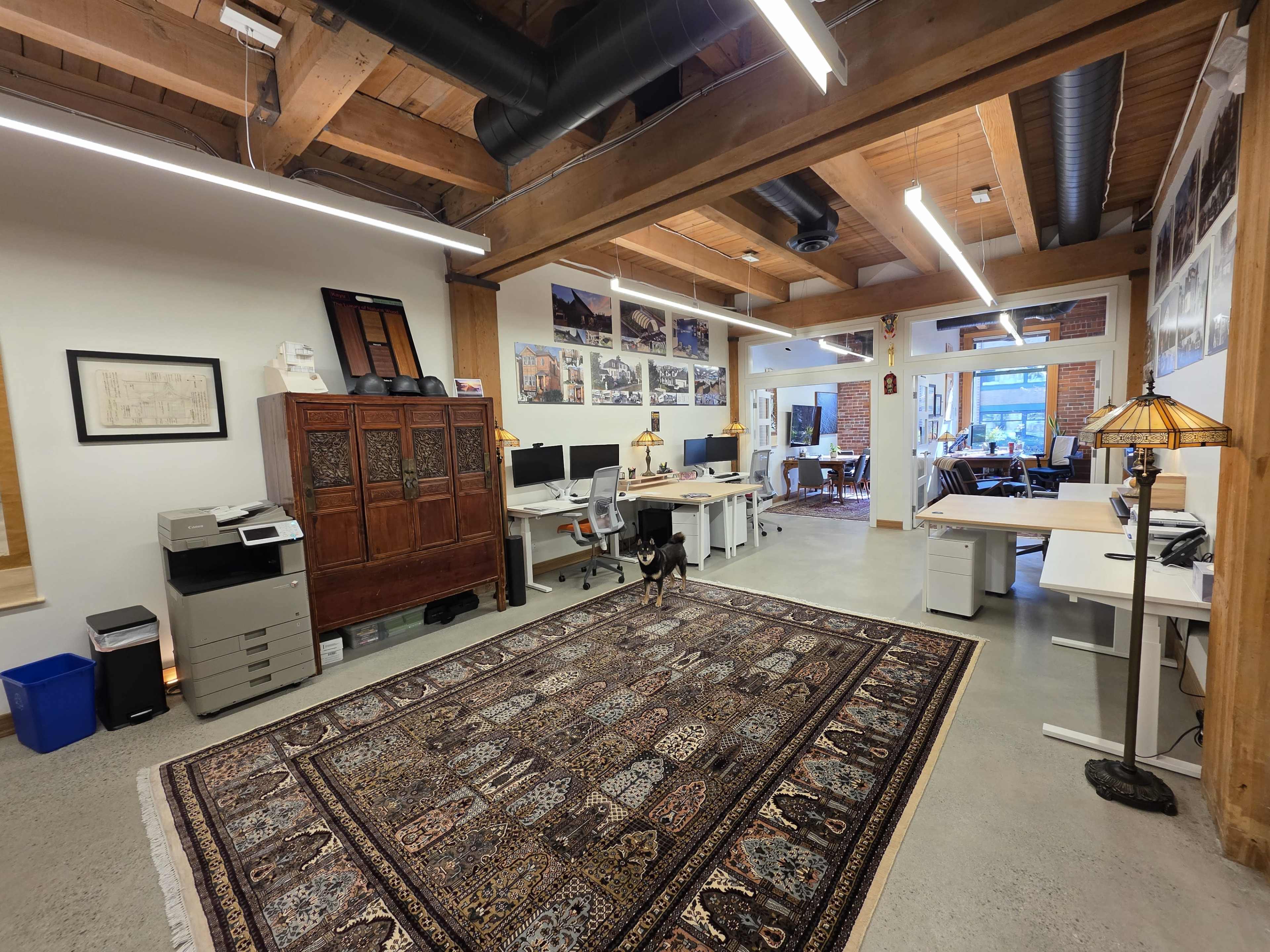 The image shows a spacious office with wooden beams, multiple workstations, a patterned rug, and a dog standing on the carpet.