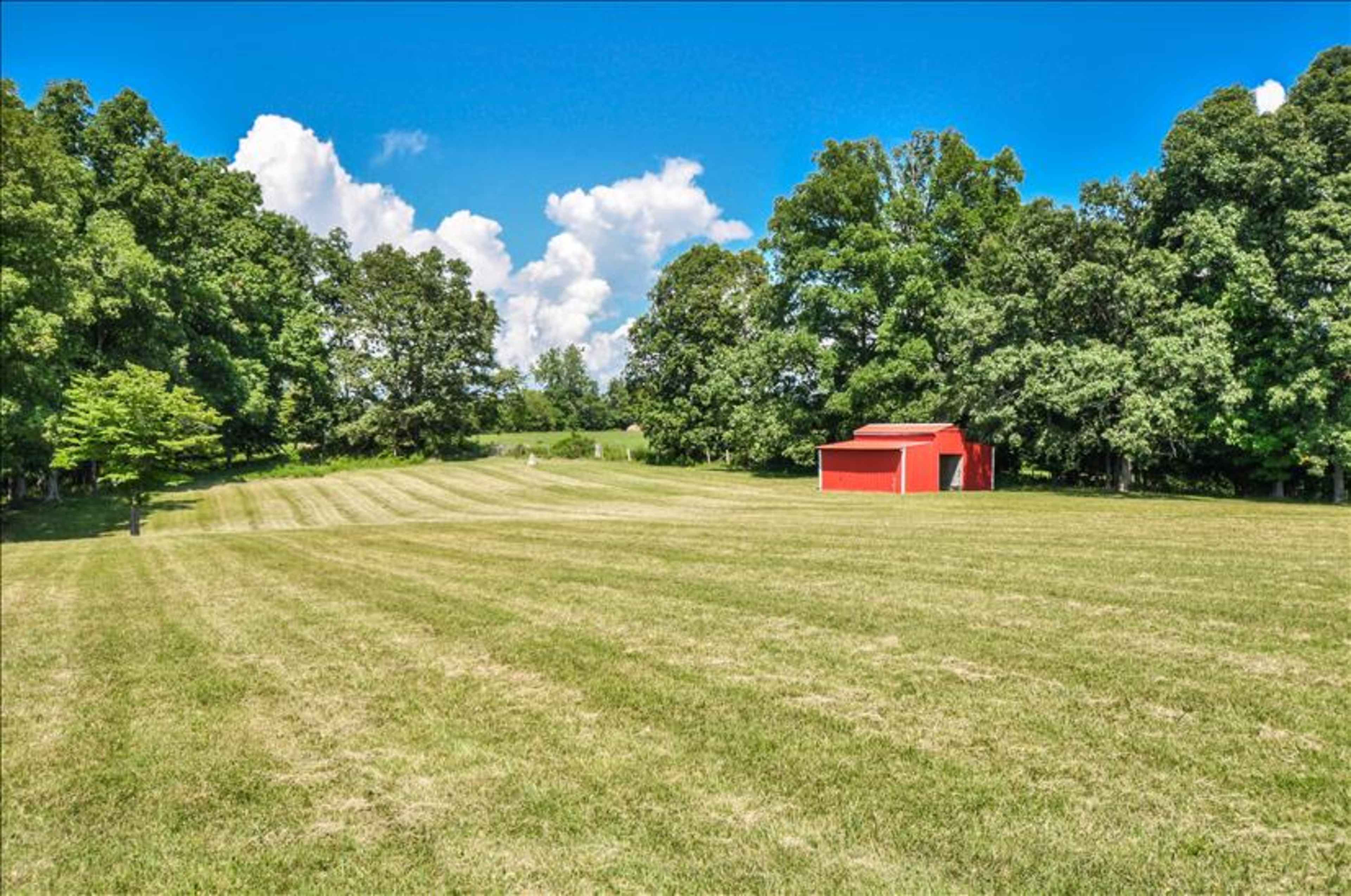 A red barn stands in a grassy field surrounded by trees under a bright blue sky.