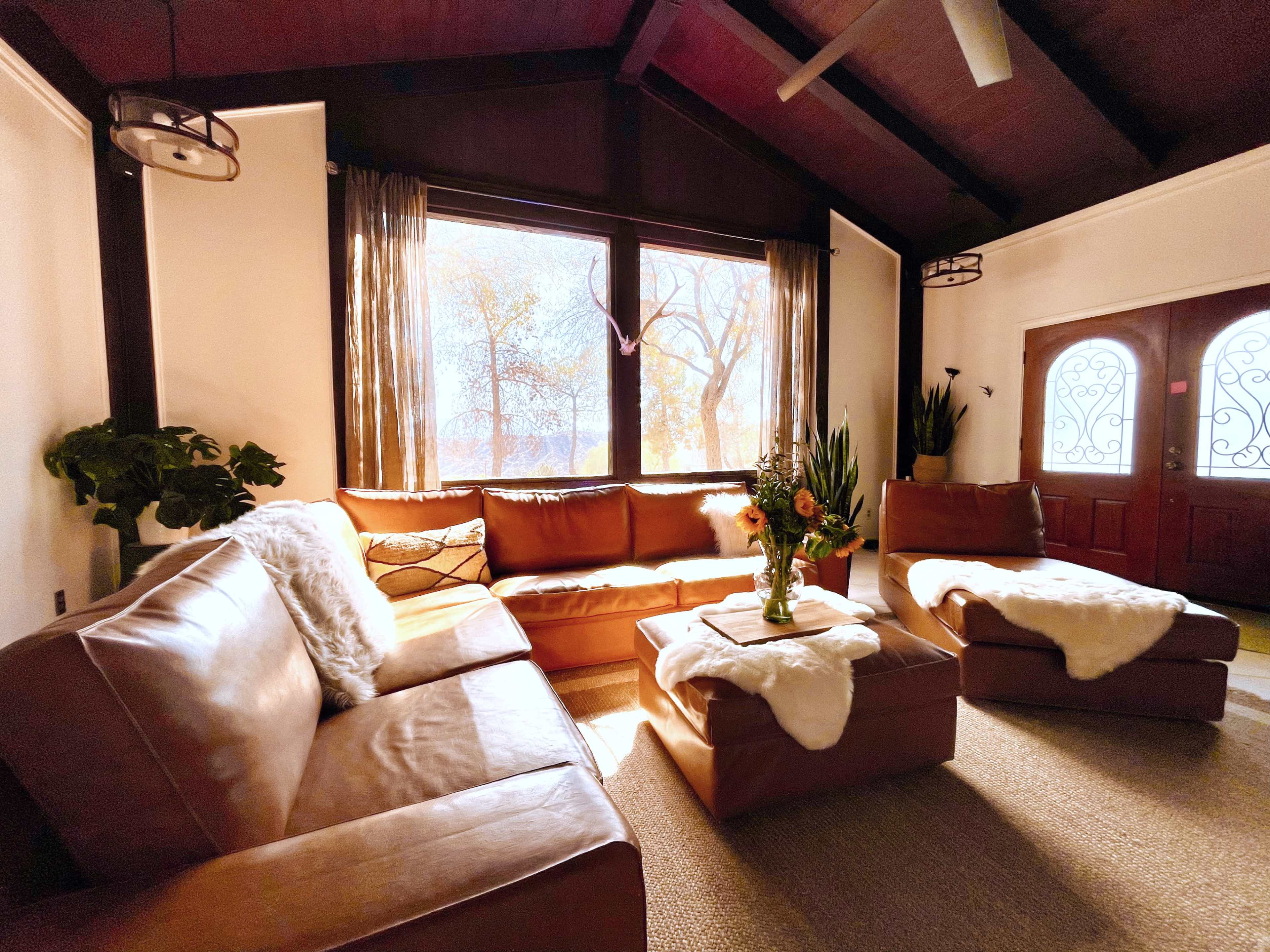 A cozy living room features a brown leather sectional sofa arranged around a coffee table with a vase of flowers, illuminated by natural light streaming through large windows.