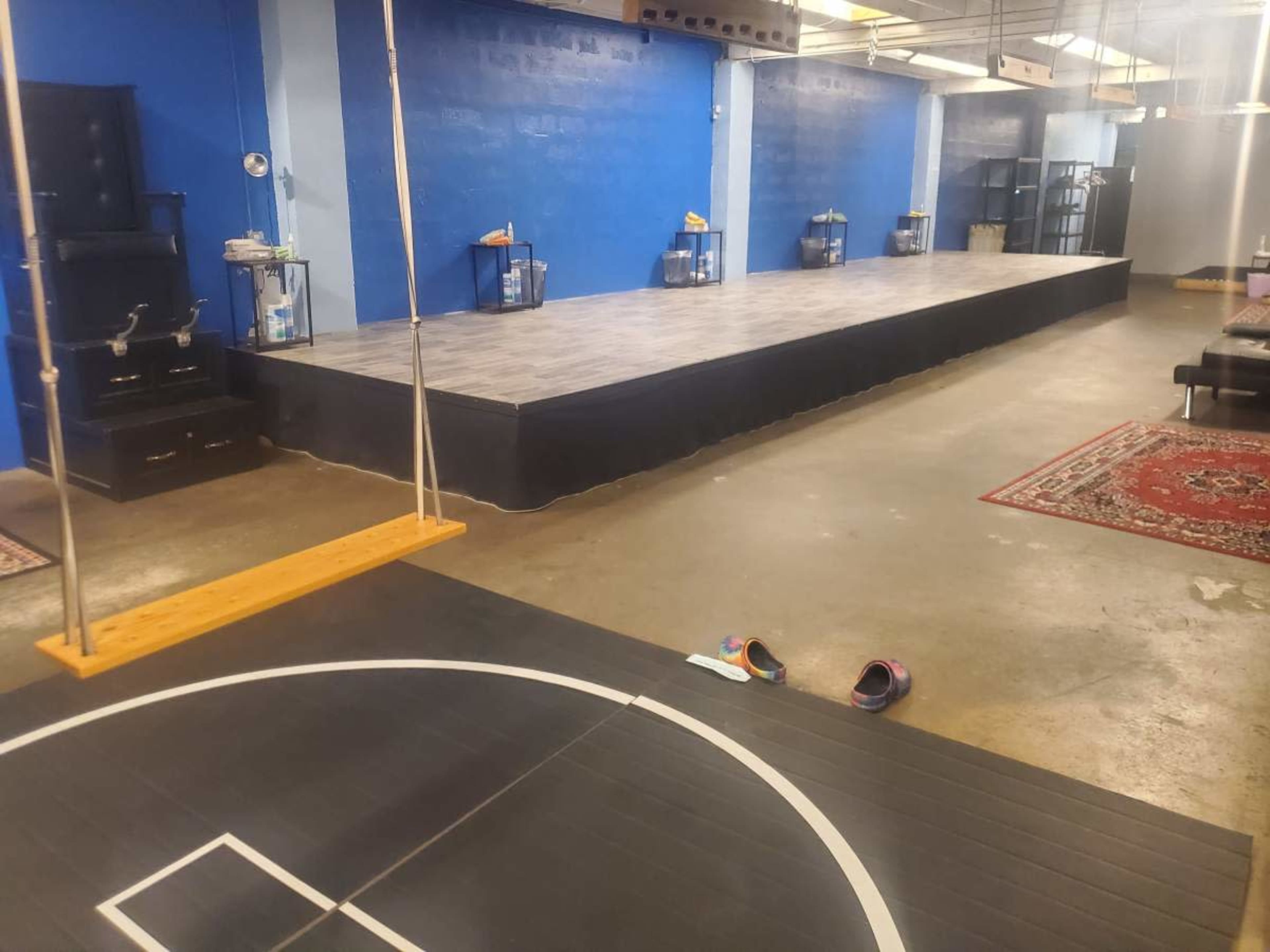 The image shows a spacious indoor area with a wooden stage, basketball court markings, and a swing in the foreground.