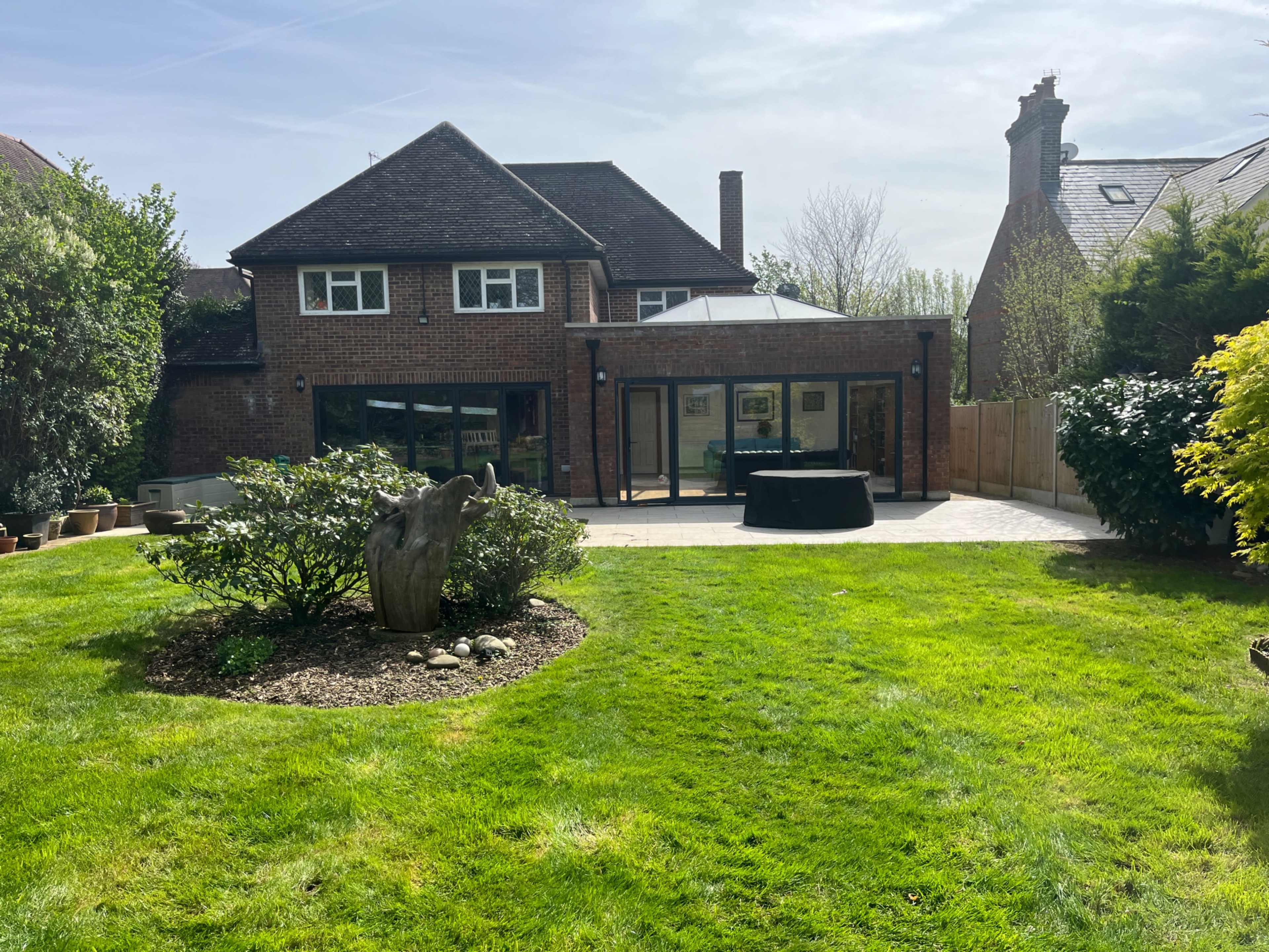 The image shows a brick house with large glass windows, a patio area, and a landscaped garden featuring grass, shrubs, and a small tree stump.