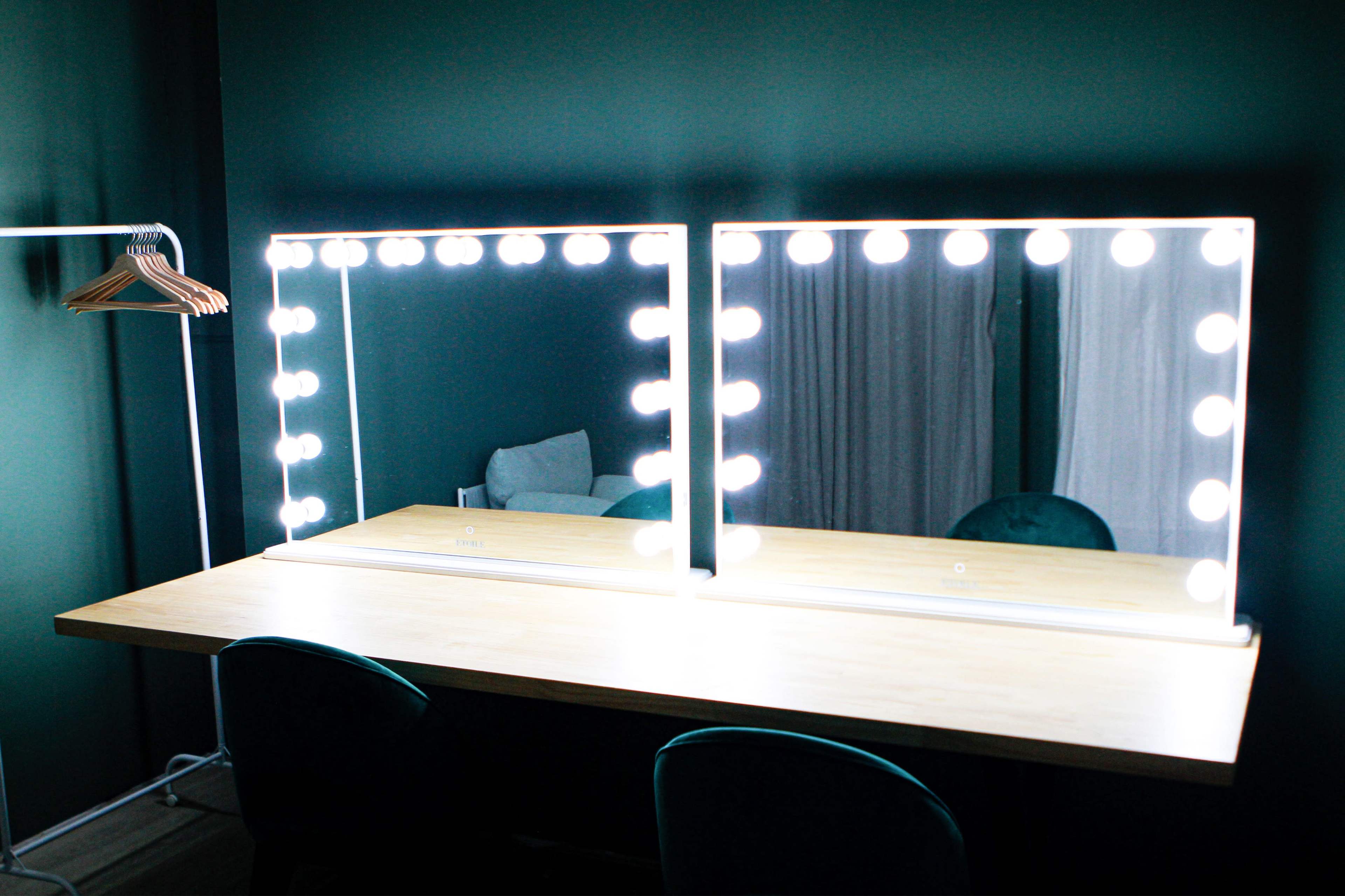 A well-lit vanity area features two mirrors surrounded by bright bulbs, set against a dark green wall, with a wooden table and a clothing hanger nearby.