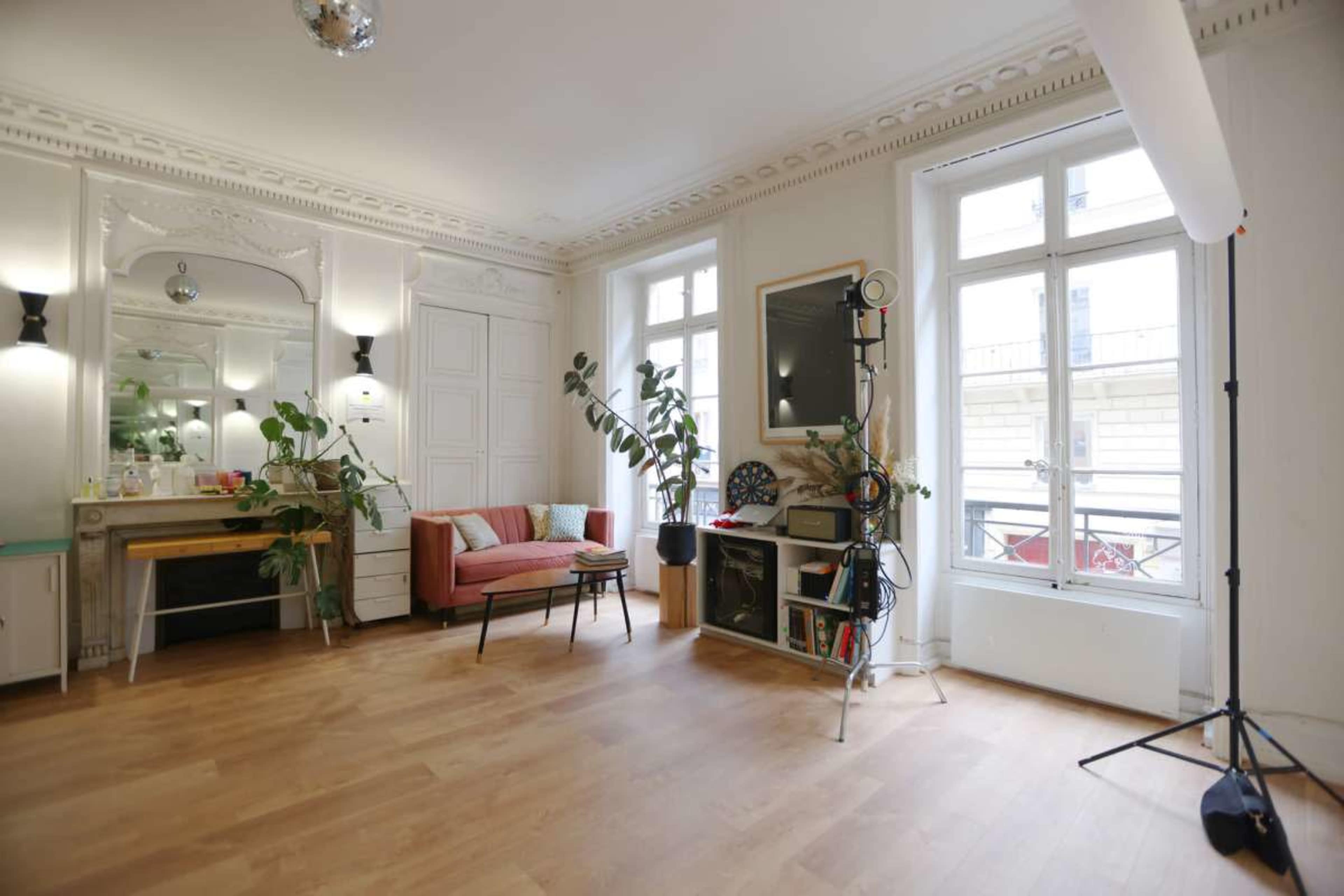 The image shows a well-lit, spacious room featuring a pink couch, wooden flooring, and a mix of decorative plants and furniture.