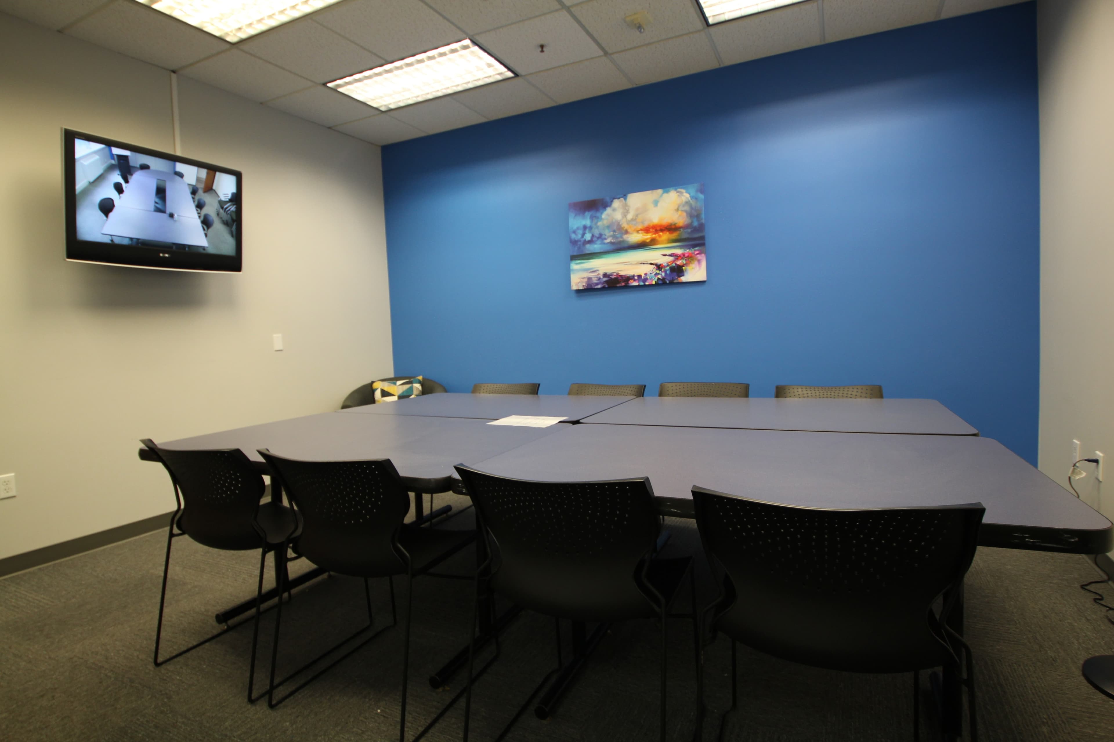 A conference room features a long table surrounded by black chairs, with a television mounted on the wall and a painting above the table.