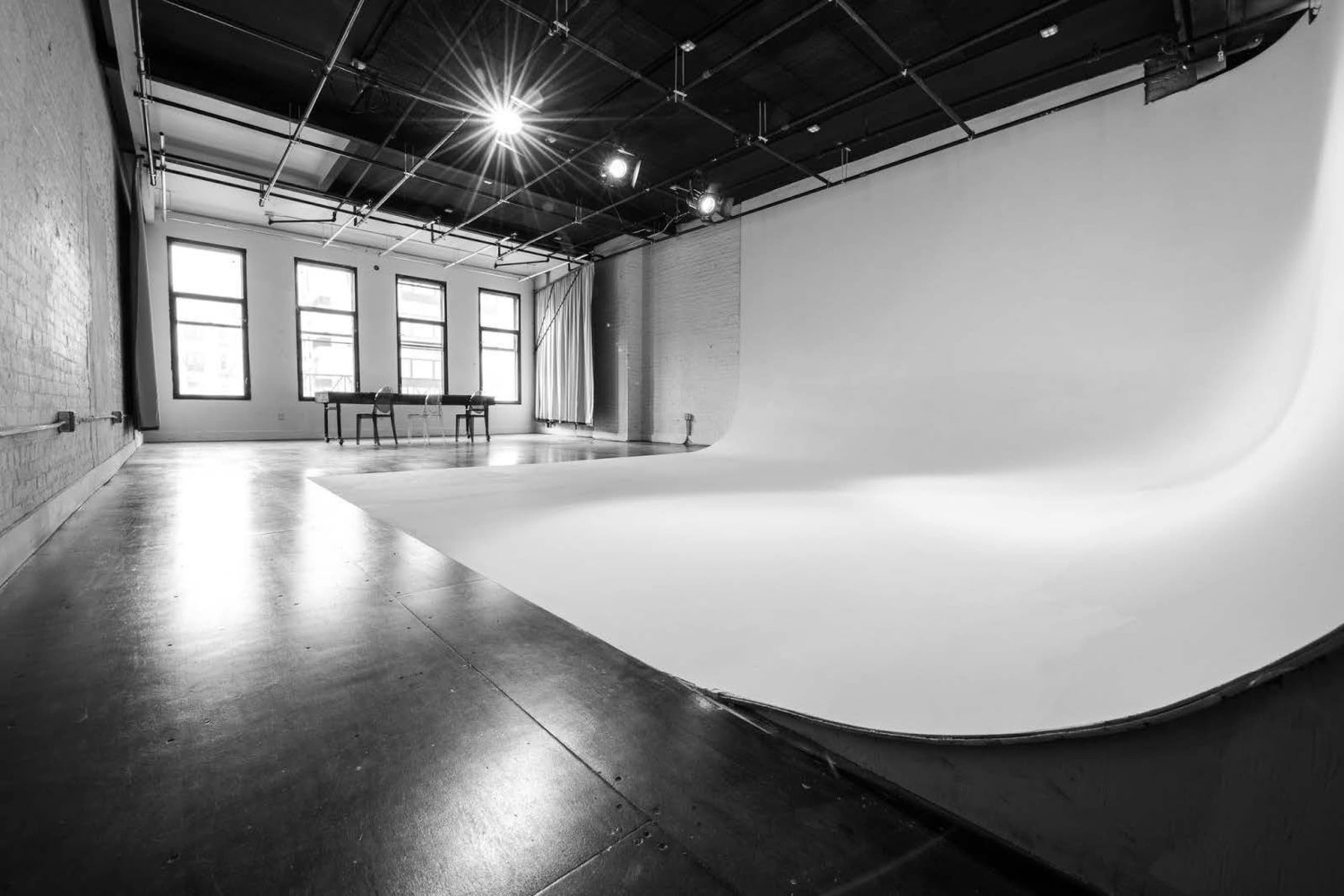 A spacious photography studio features a seamless white backdrop, large windows, and a simple table in the background.