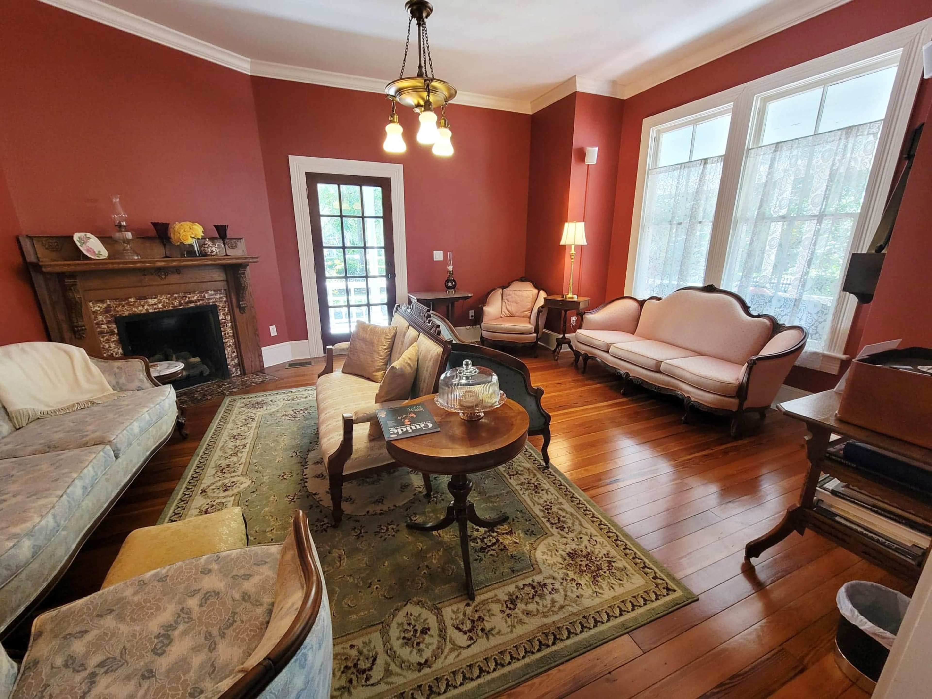 The room features hardwood floors, a patterned rug, vintage furniture, and large windows that provide natural light.