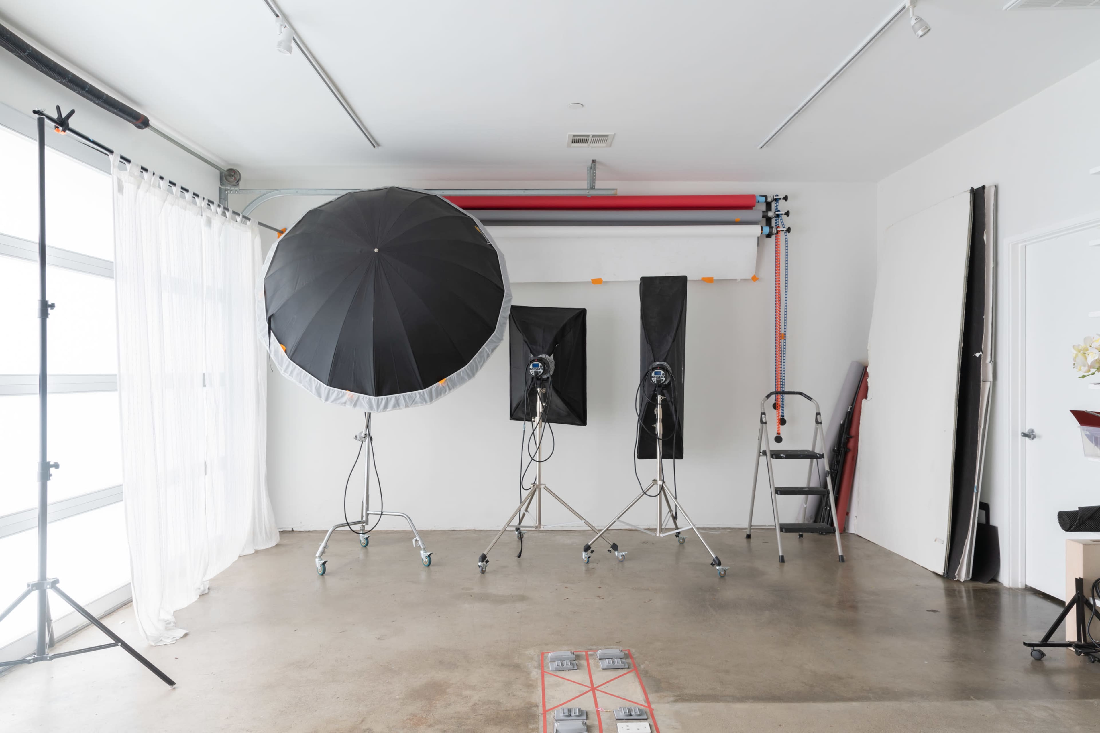 A photography studio with two large light umbrellas, two softboxes on stands, a ladder, and a backdrop setup against a white wall.