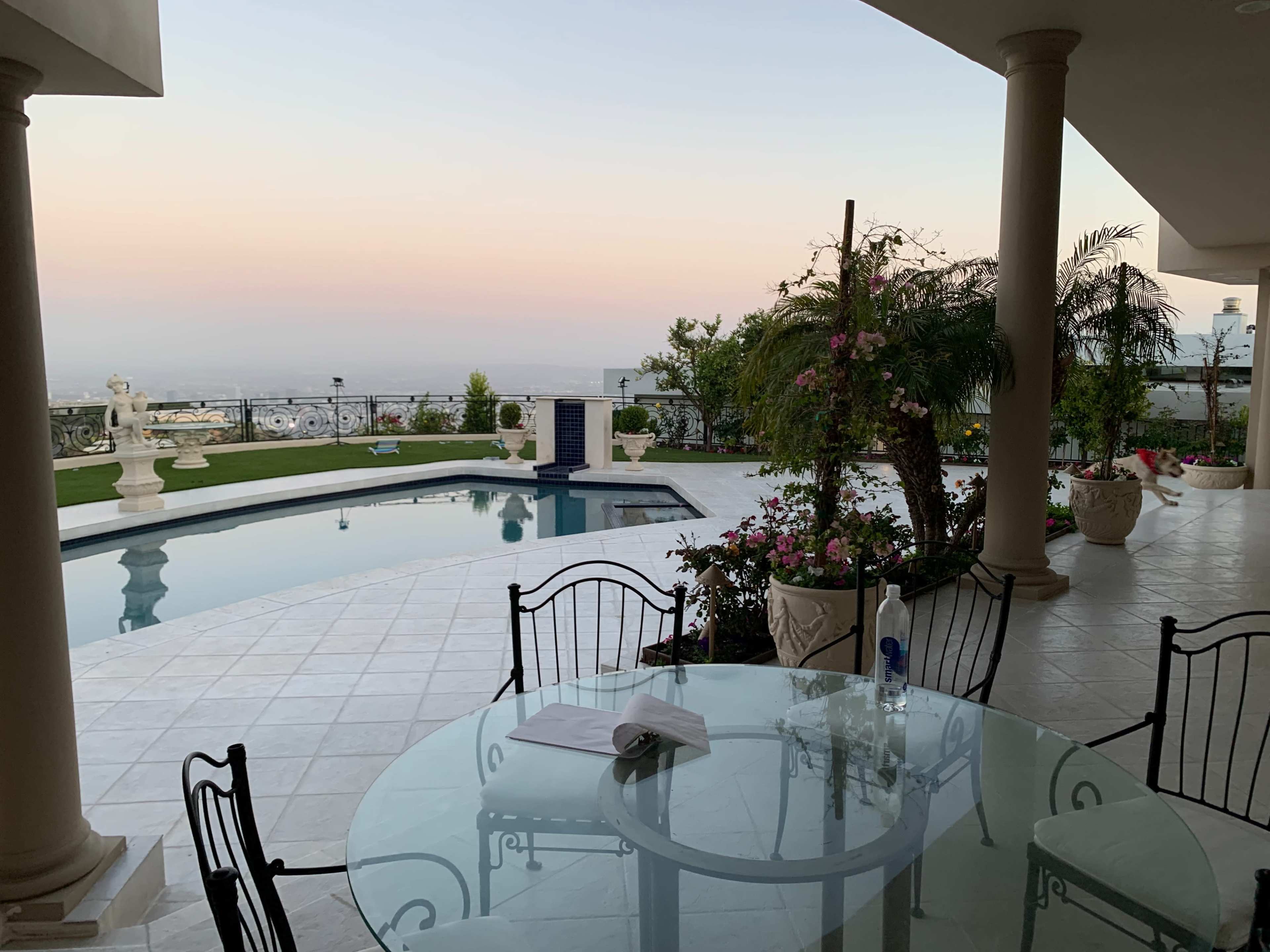 A patio area with a glass table and chairs overlooks a swimming pool surrounded by gardens and a clear sky at dusk.