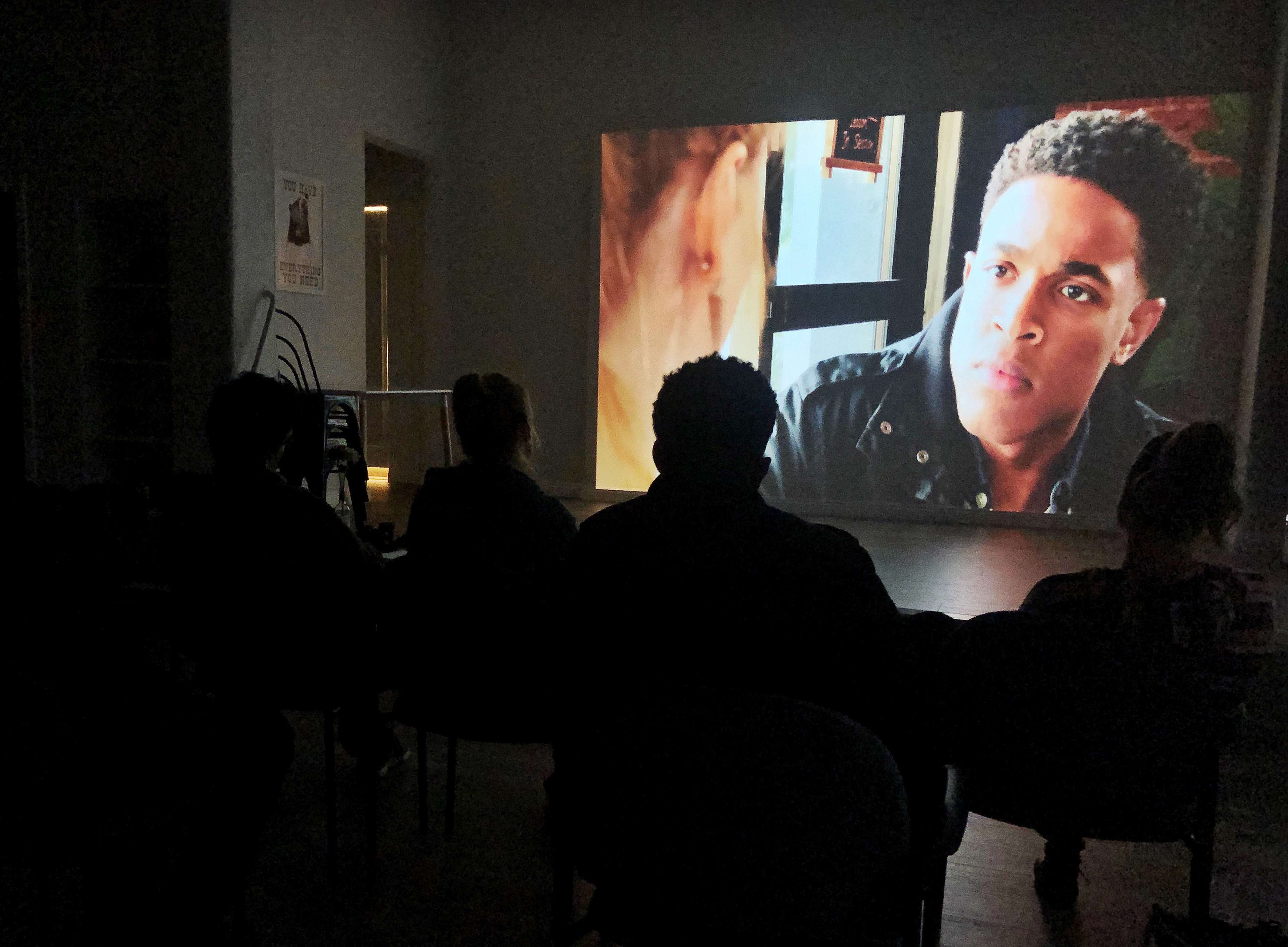 A group of people watches a movie projected on a wall in a dimly lit room.