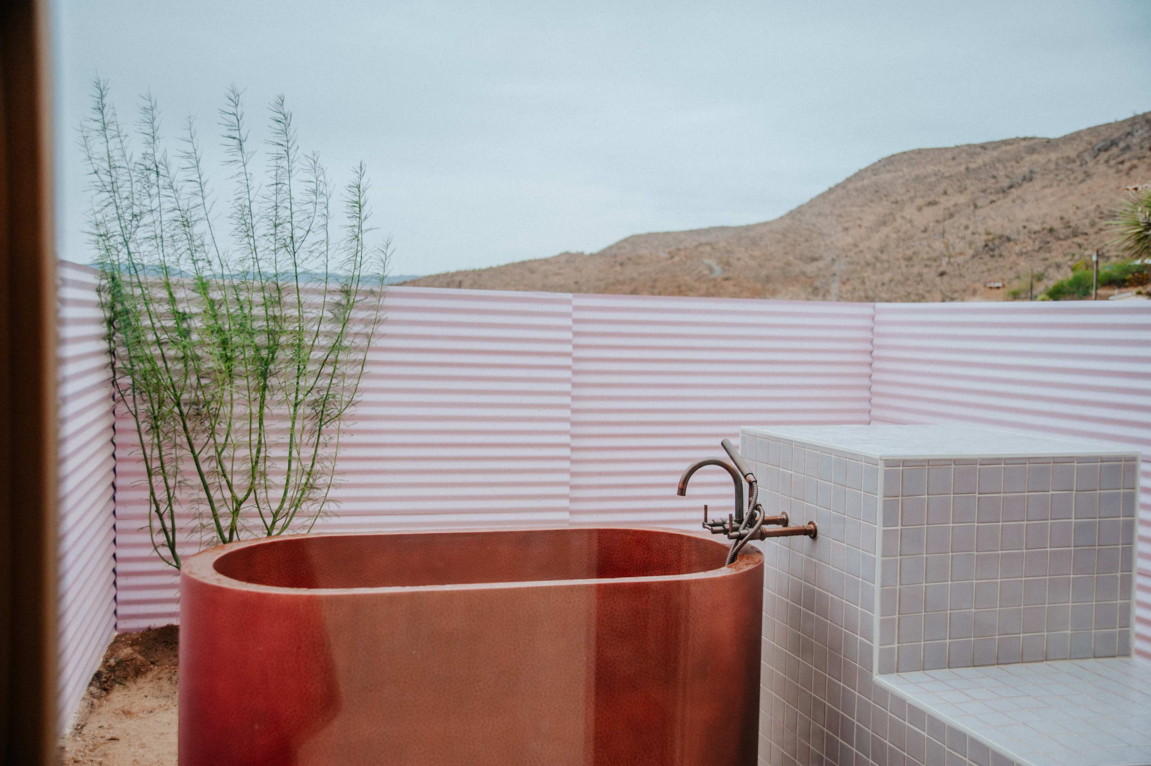 A stylish outdoor bathtub made of metal, surrounded by a pink corrugated fence, with a desert landscape in the background.