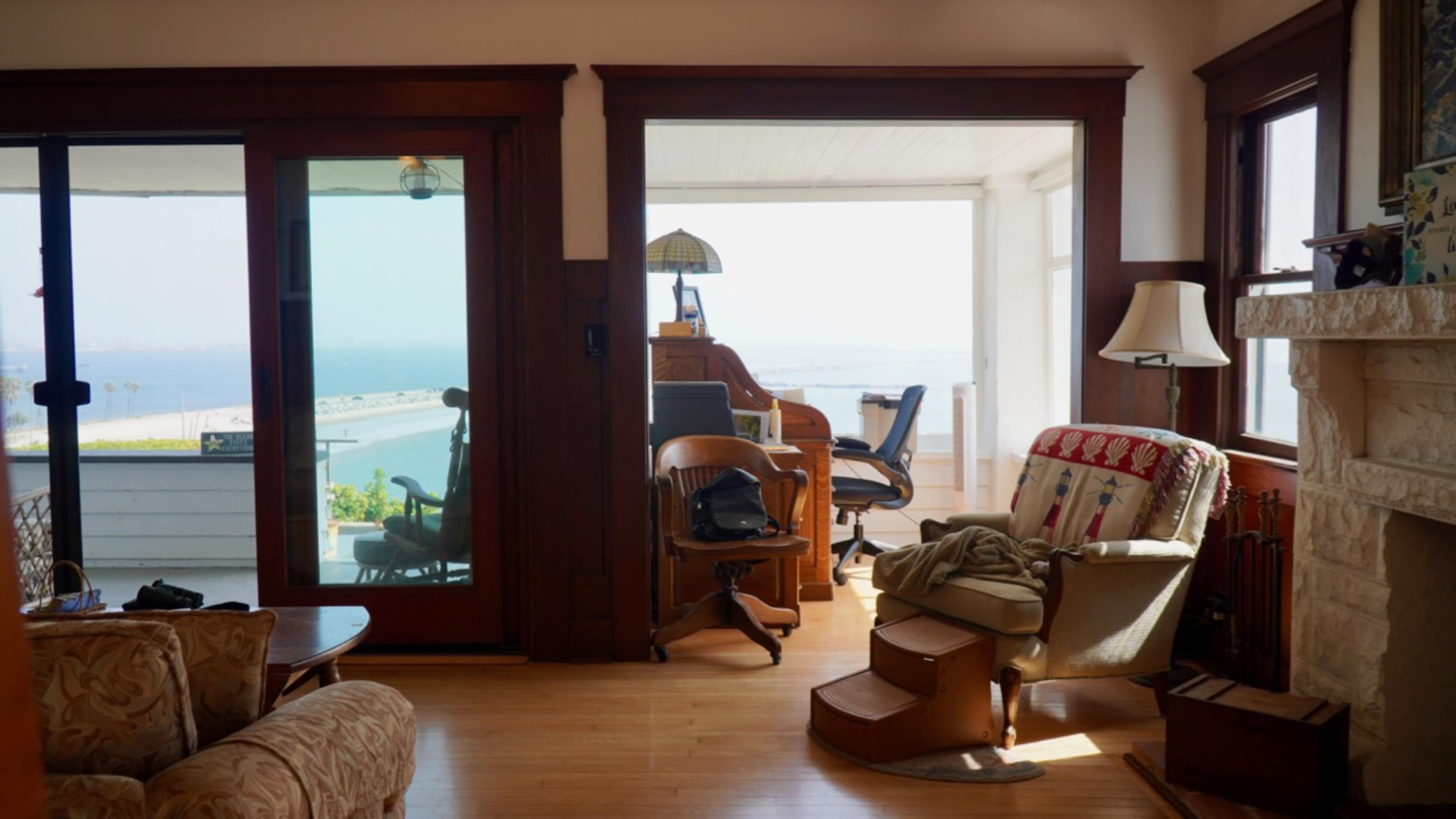 The image shows a room with wooden accents, featuring a view of the ocean through large glass doors, a cozy seating area, and a workspace with a chair and desk.