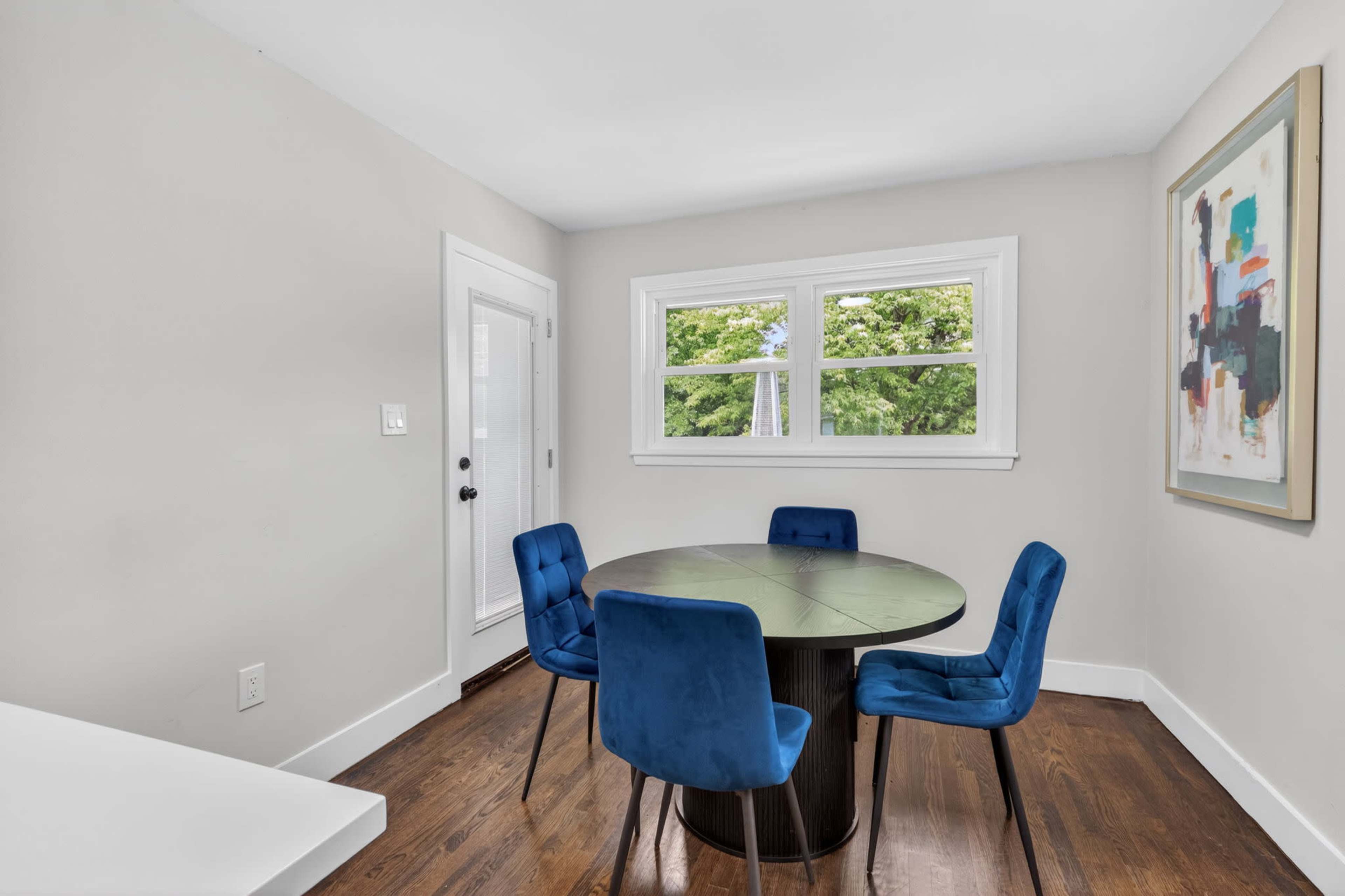 A round table with four blue velvet chairs is positioned in a room featuring large windows and a piece of framed artwork on the wall.