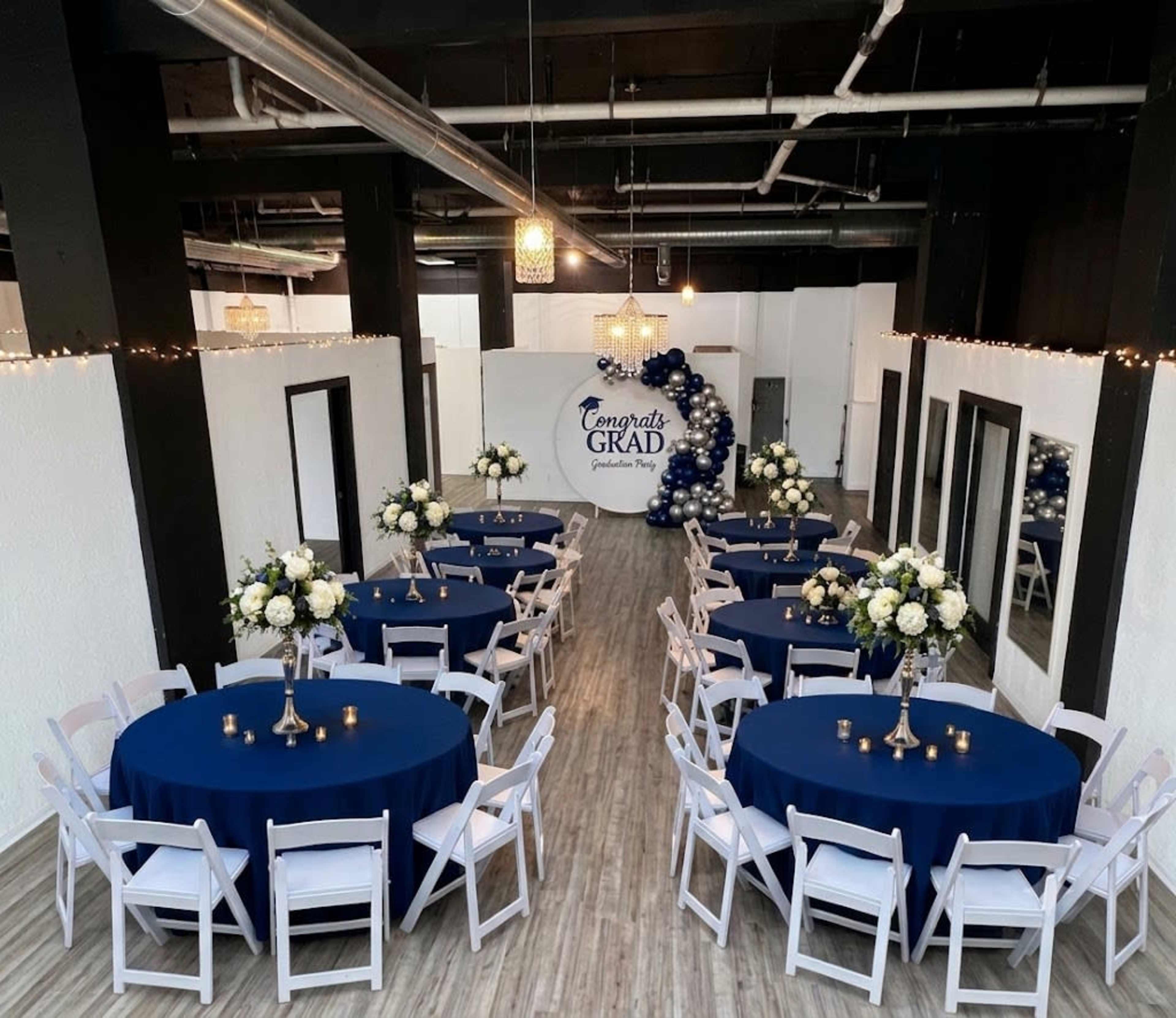 The image shows a decorated event space with round tables covered in navy blue tablecloths, floral centerpieces, and a backdrop reading "Congrats Grad" surrounded by balloons.