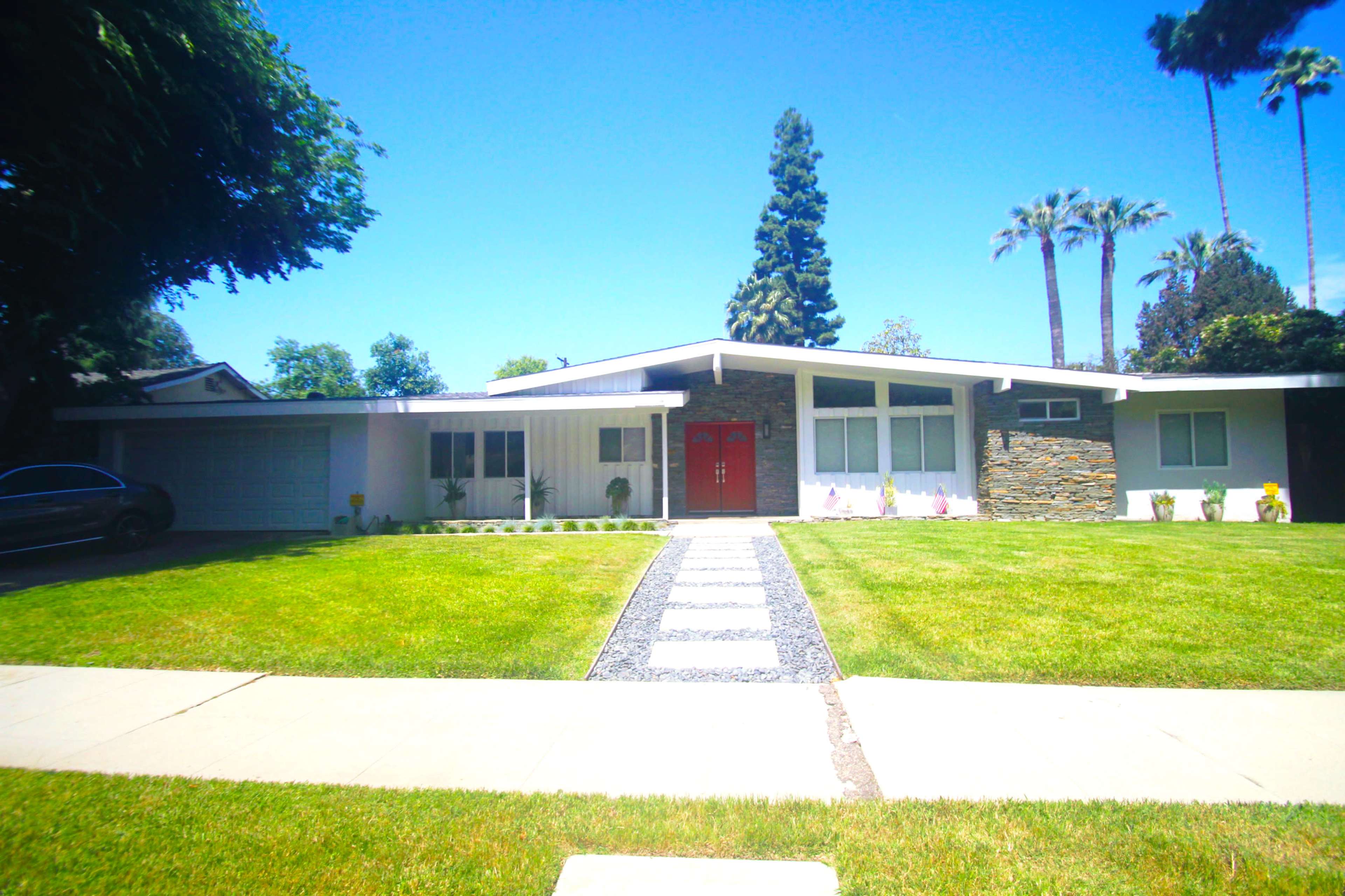 A modern single-story house features a red double door, stone accents, and a neatly paved path leading to the entrance, surrounded by a manicured lawn and palm trees.