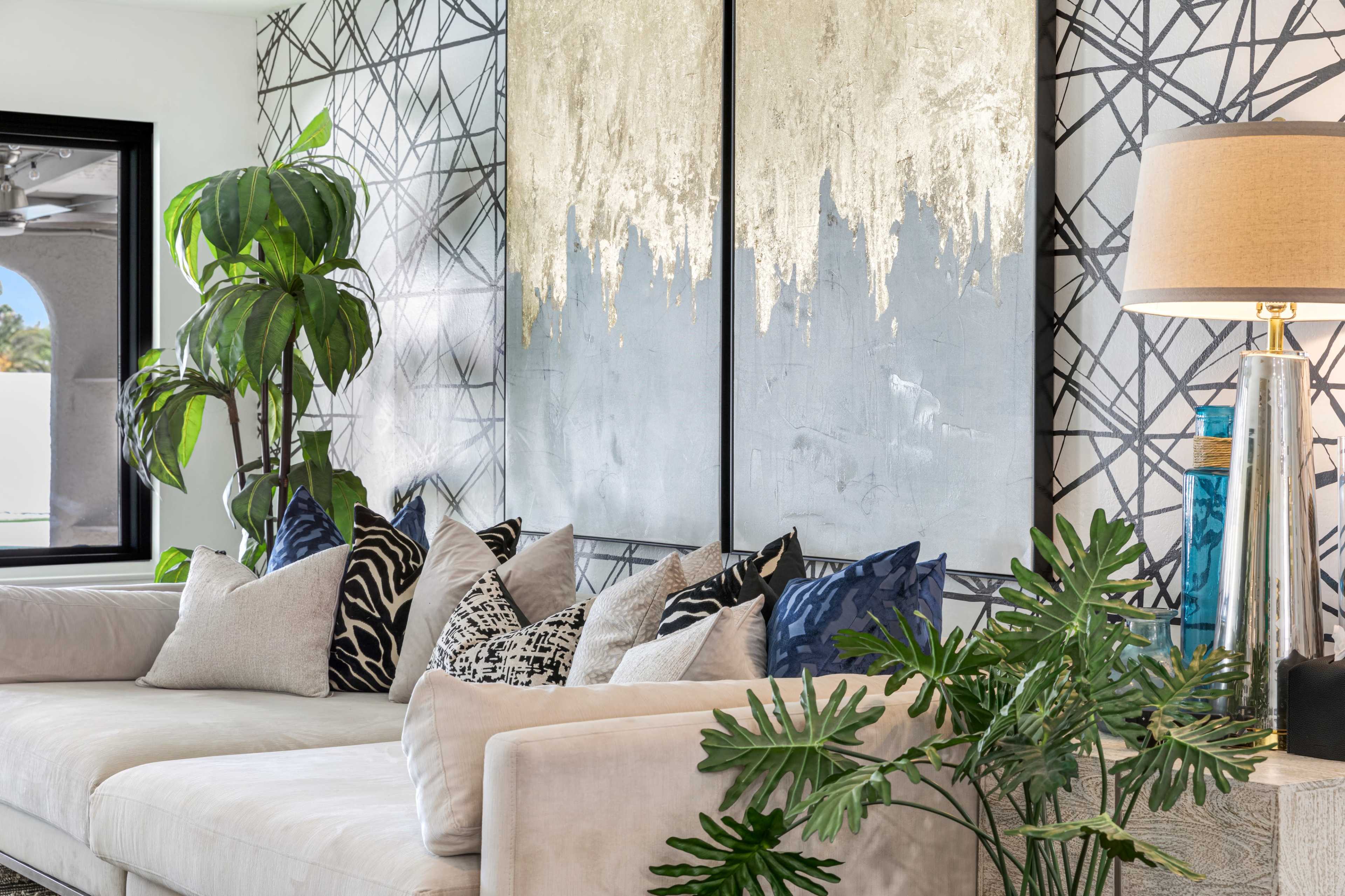 A modern living room features a light sofa, decorative pillows, a large abstract painting, and indoor plants beside a lamp.