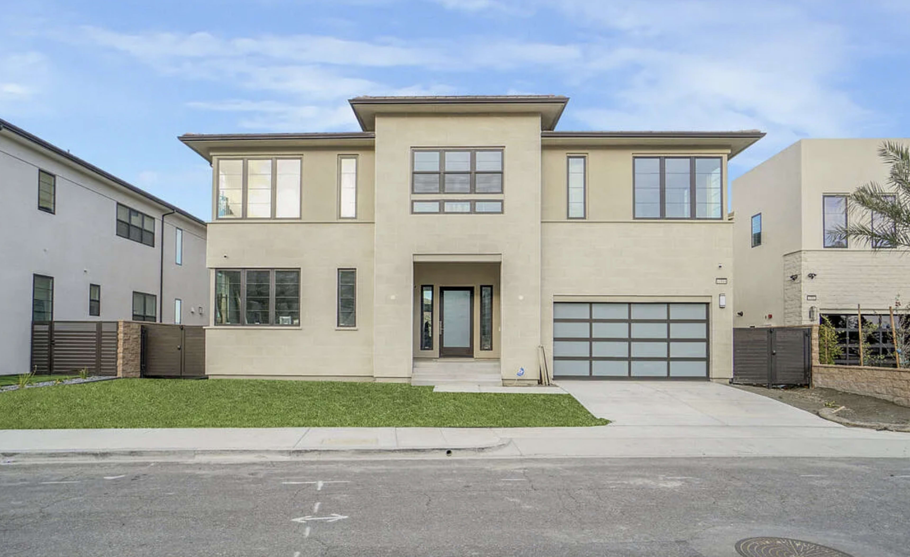 The image shows a modern, two-story house with a beige exterior, multiple windows, and a two-car garage, situated on a suburban street.