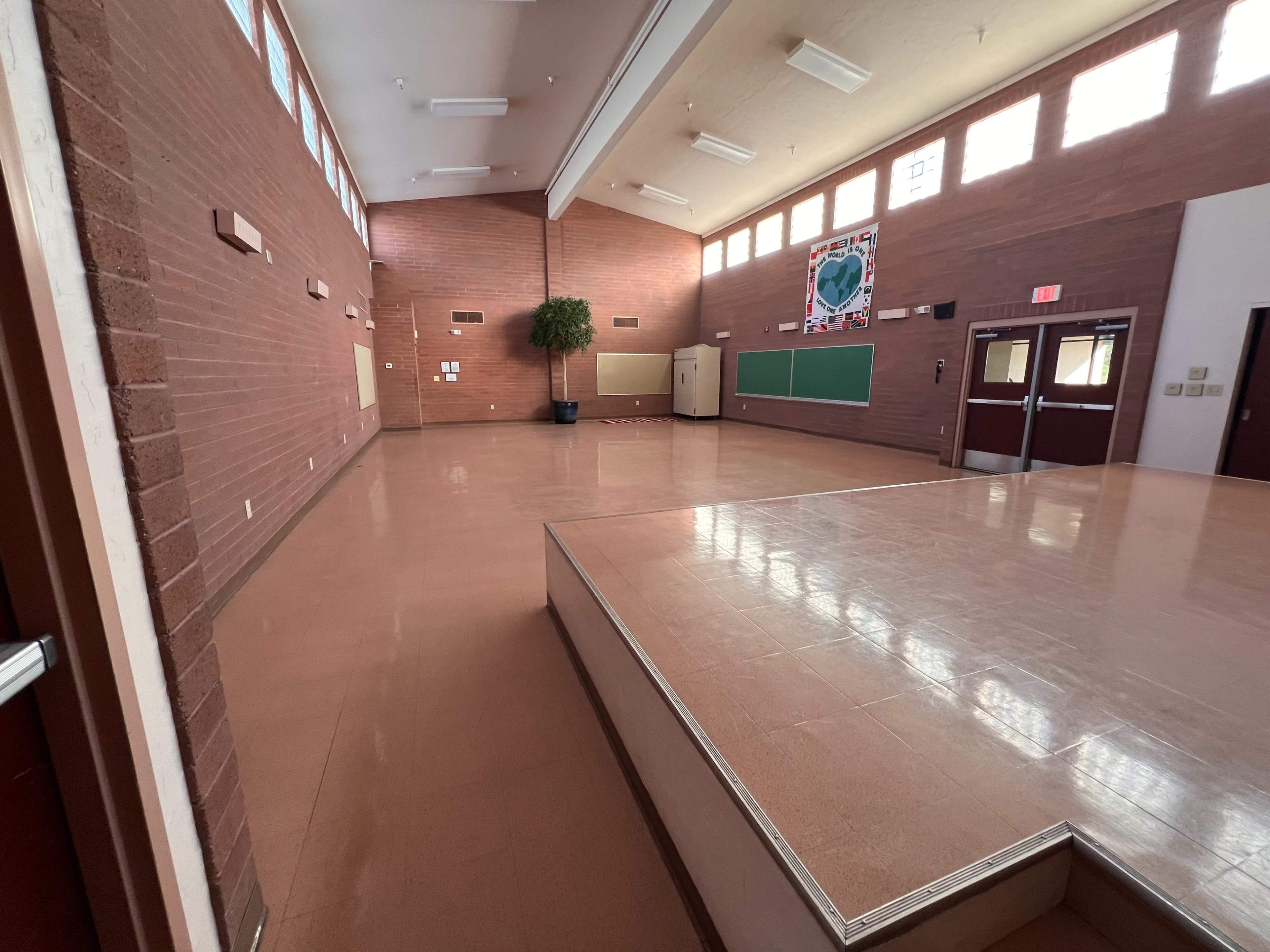 The image shows an empty multipurpose room with brown brick walls, a wooden stage, and large windows allowing natural light.