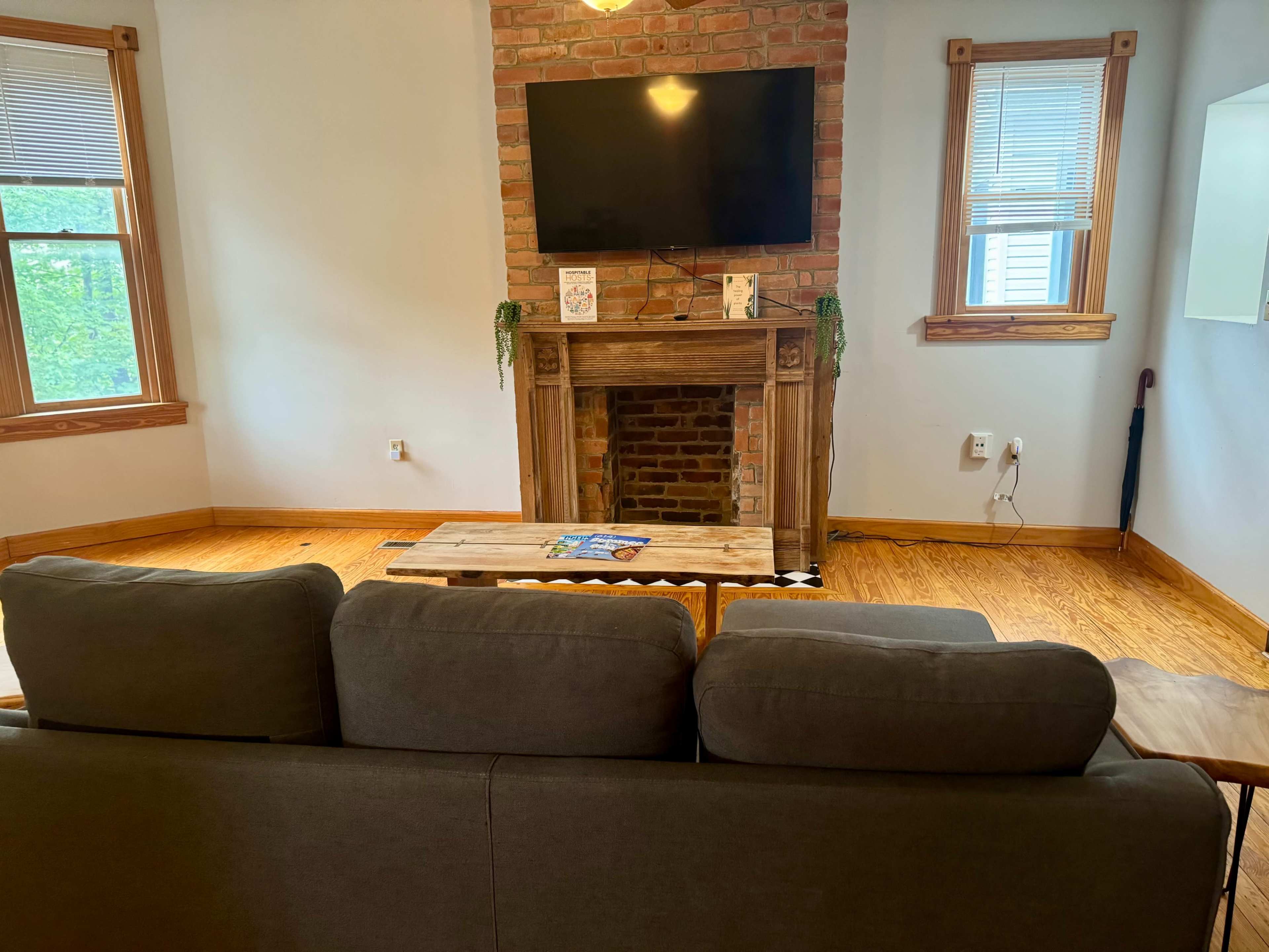 A living room features a brick fireplace with a mounted television above it, a wooden coffee table in front of a gray sofa, and large windows with natural light.
