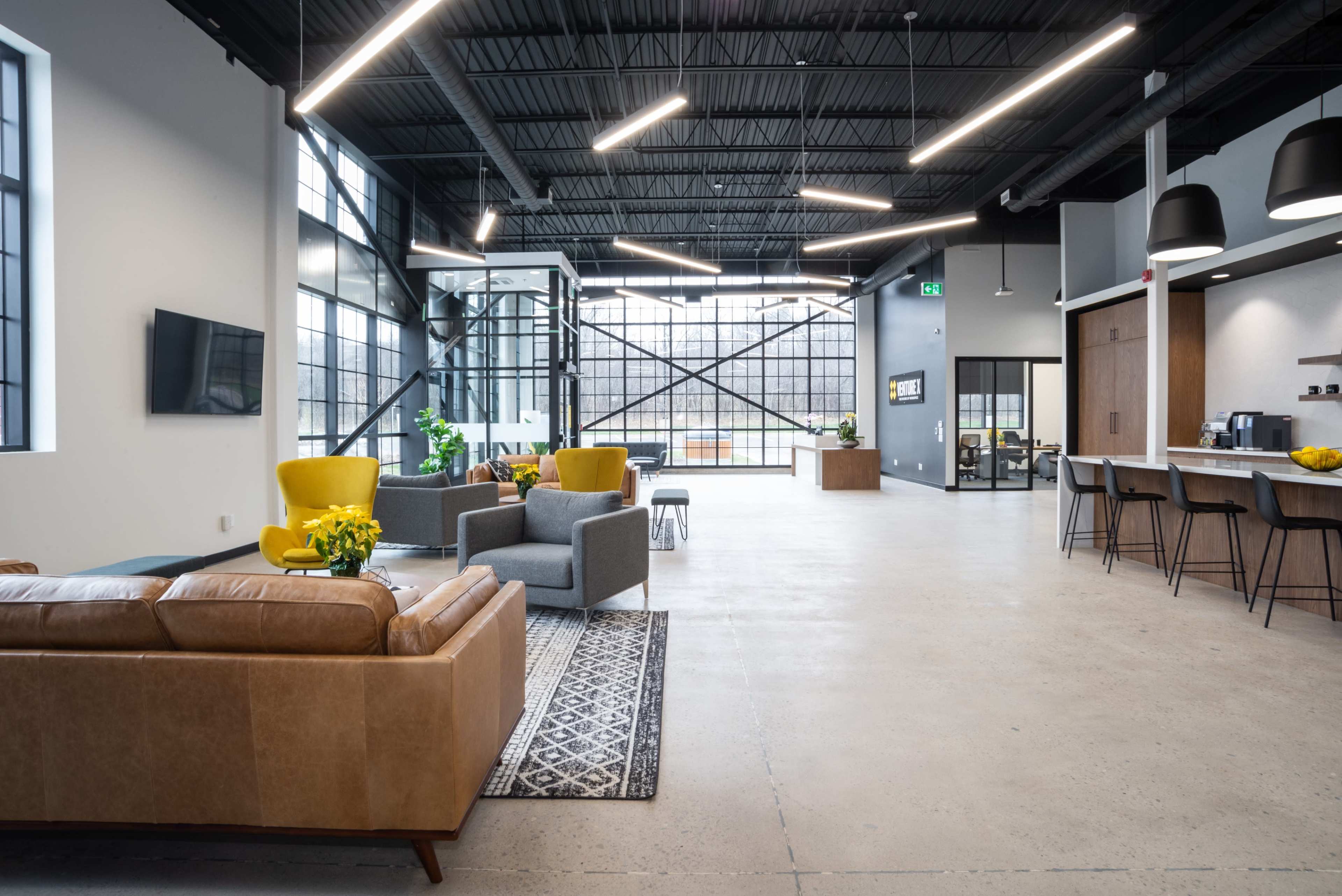 A spacious, modern lounge area featuring a leather sofa, colorful armchairs, and large windows, with a kitchen and office space visible in the background.