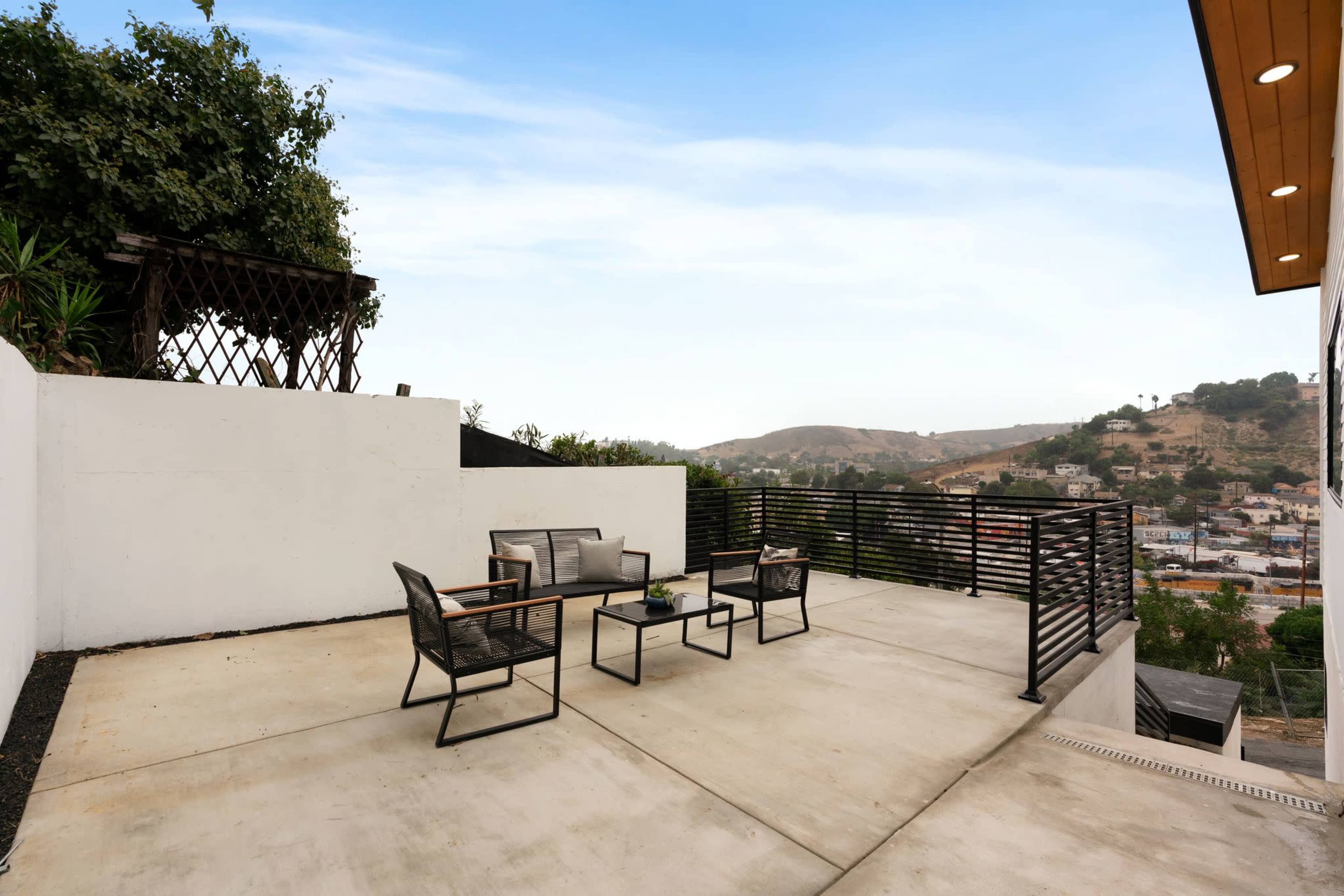 The image shows a spacious outdoor terrace with metal furniture and a view of a hilly landscape.