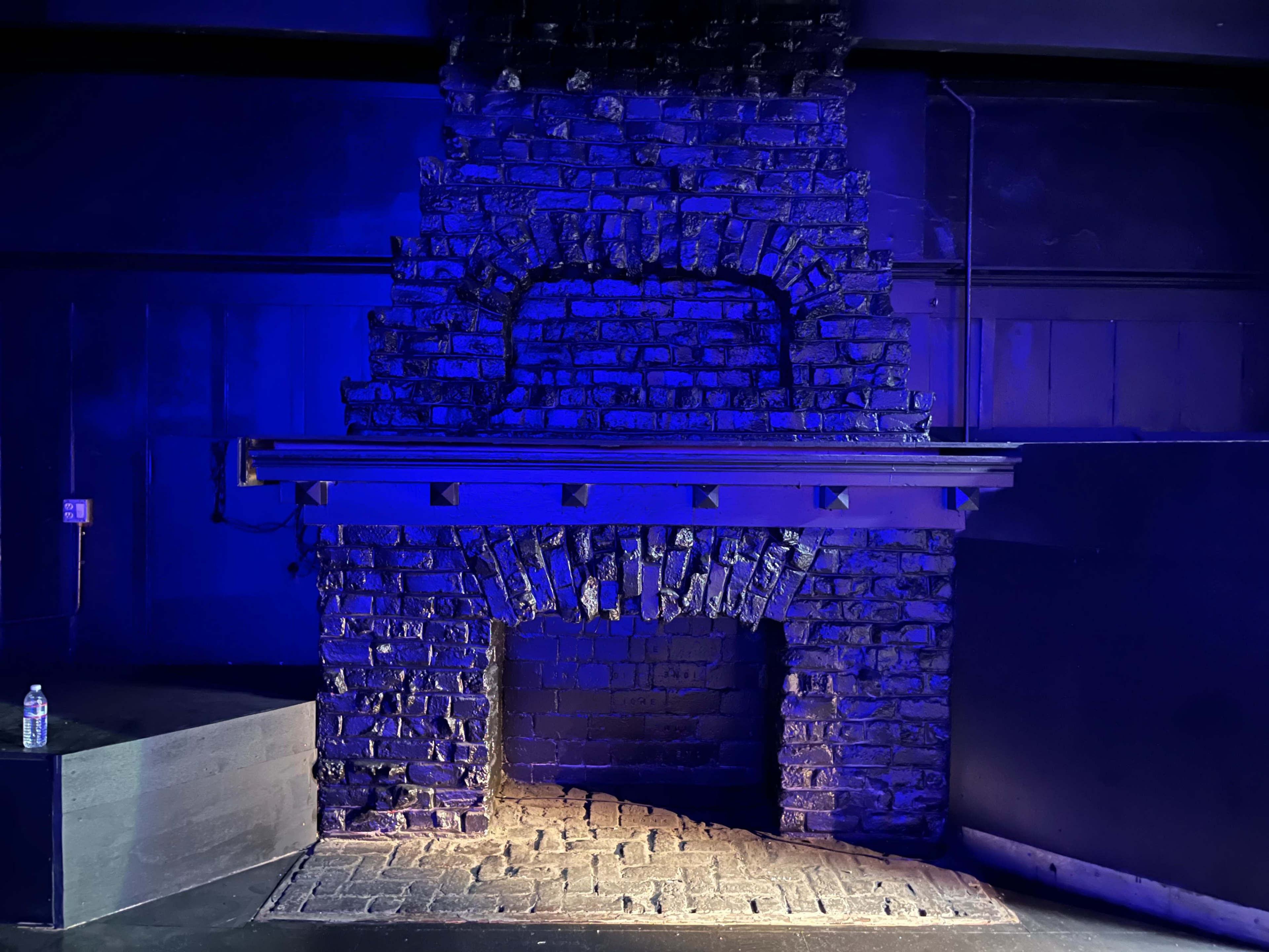 A brick fireplace with a dark blue background and a wooden mantle above it, set in an empty space with a stone floor.