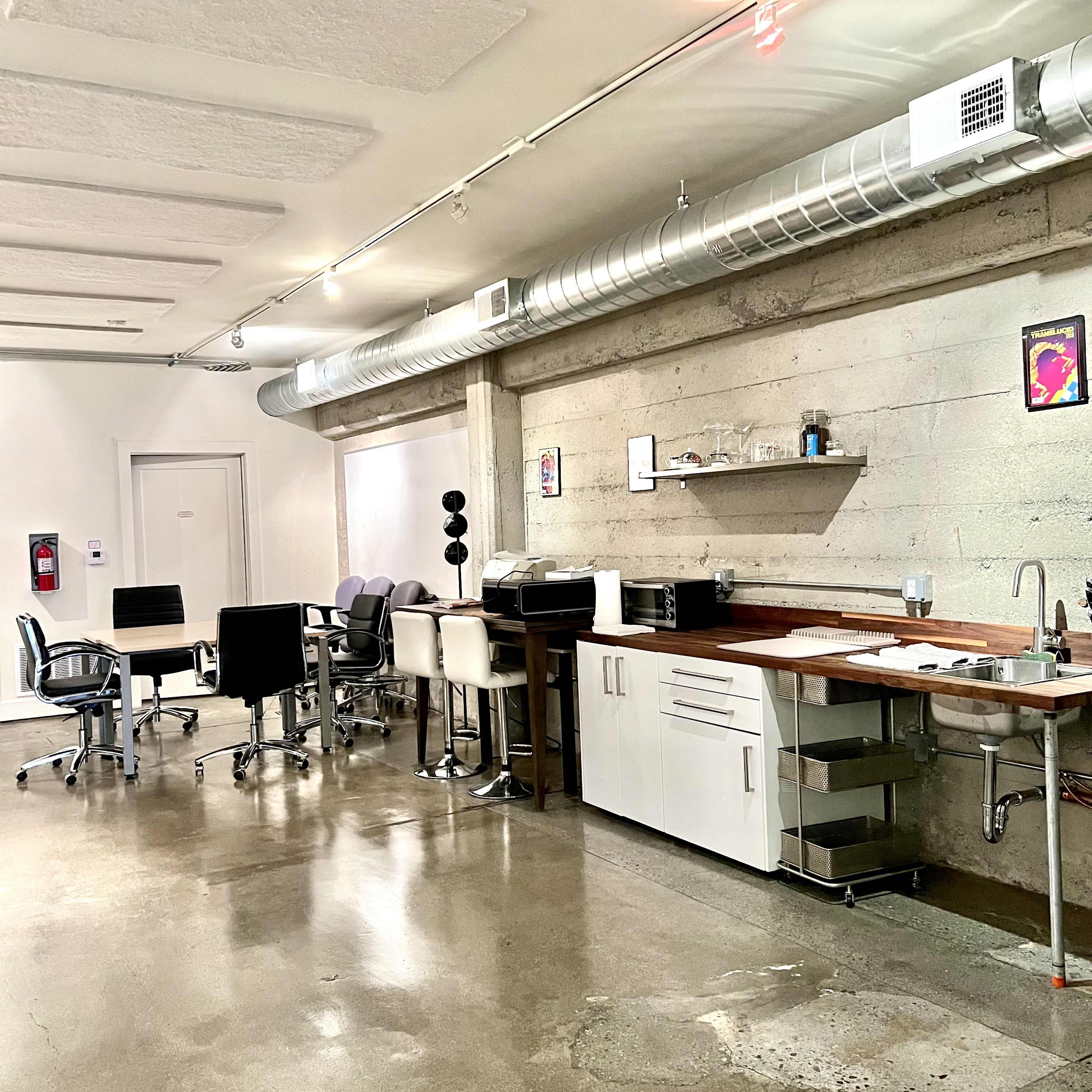A modern office space with a kitchenette on one side and several chairs and tables arranged in the area.