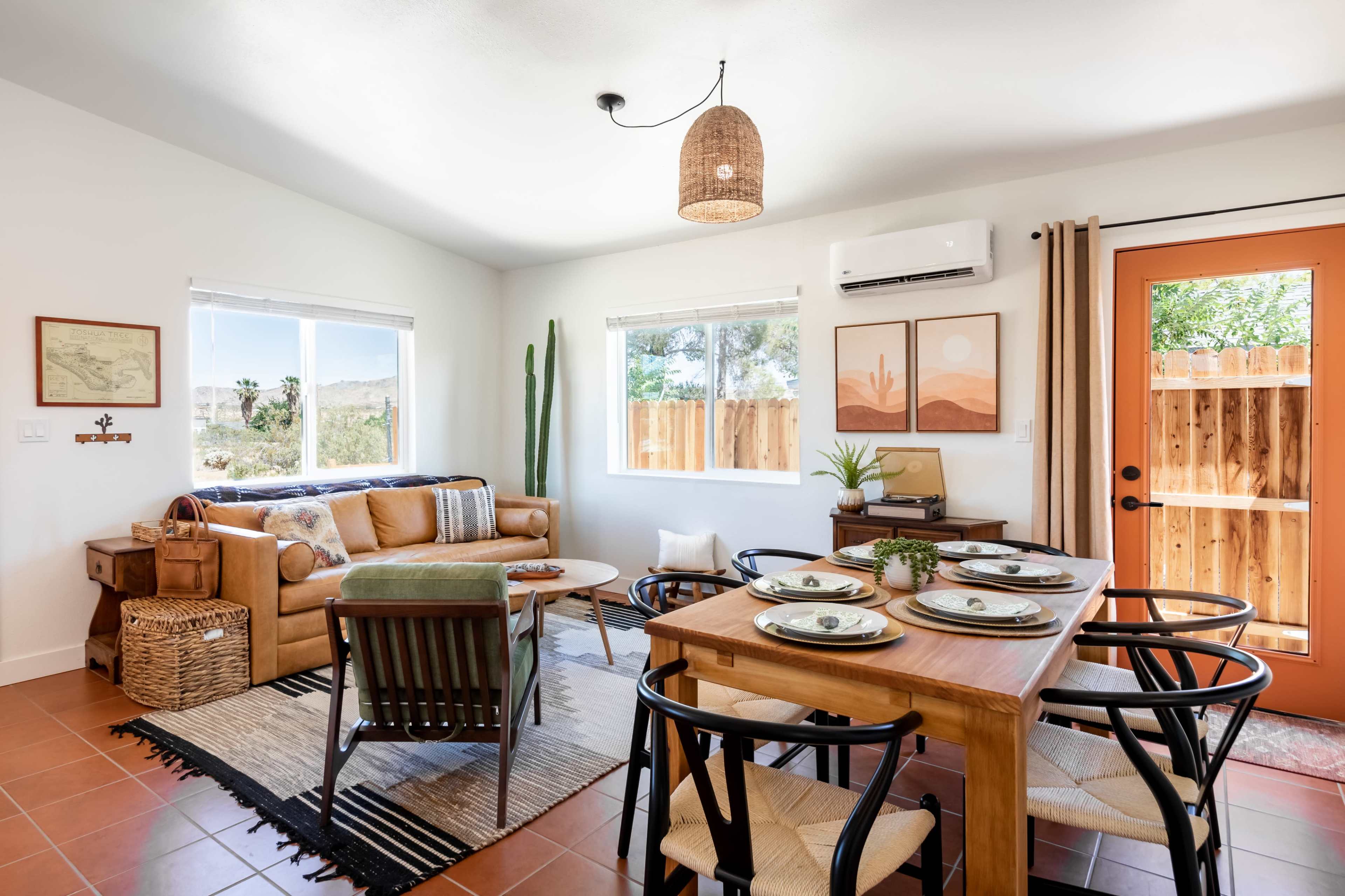 A living area with a tan couch, a wooden dining table with black chairs, and large windows allowing natural light, complemented by desert-themed artwork and a cactus.