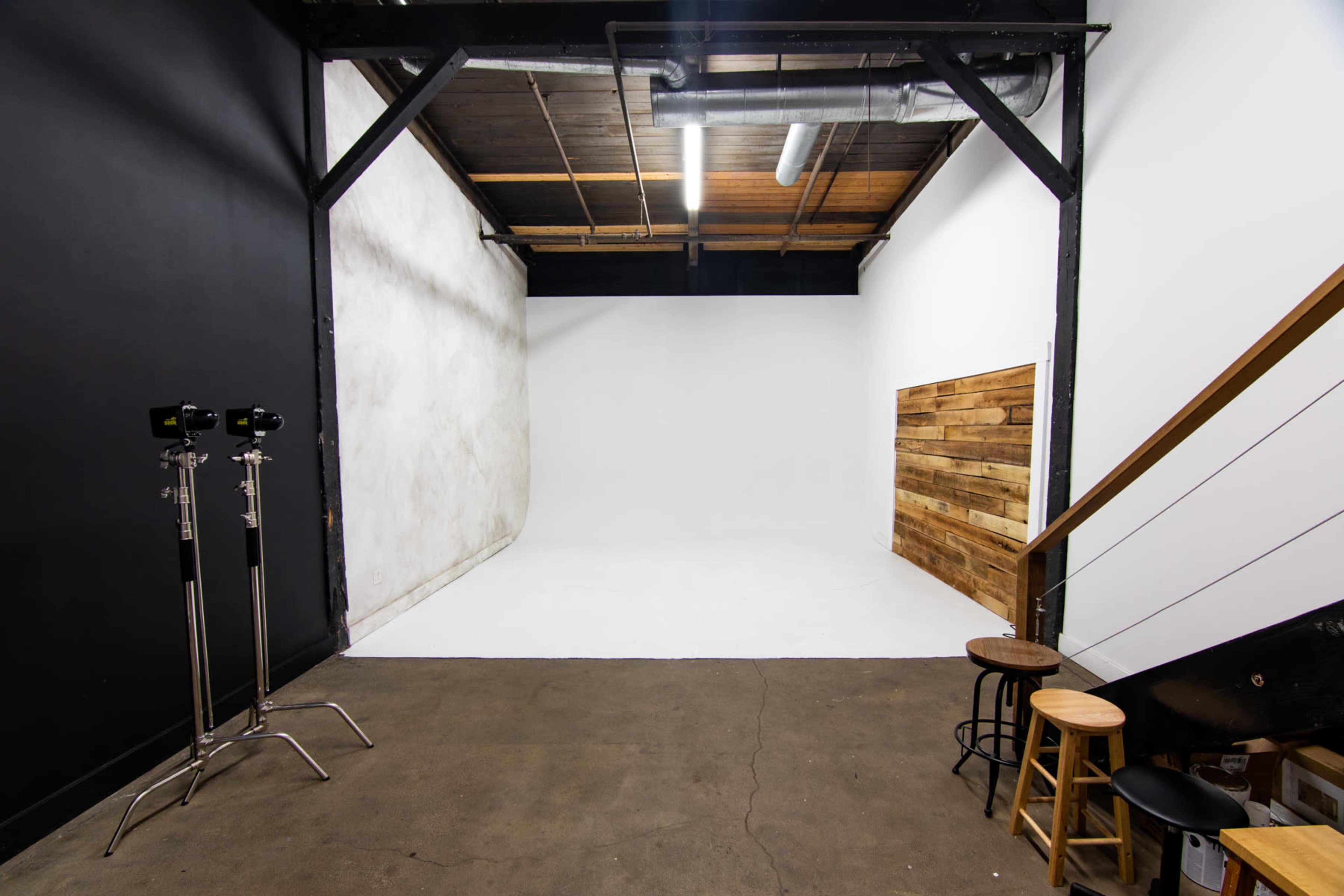 A spacious photography studio with a white backdrop, wooden accent wall, and two light stands beside a staircase.