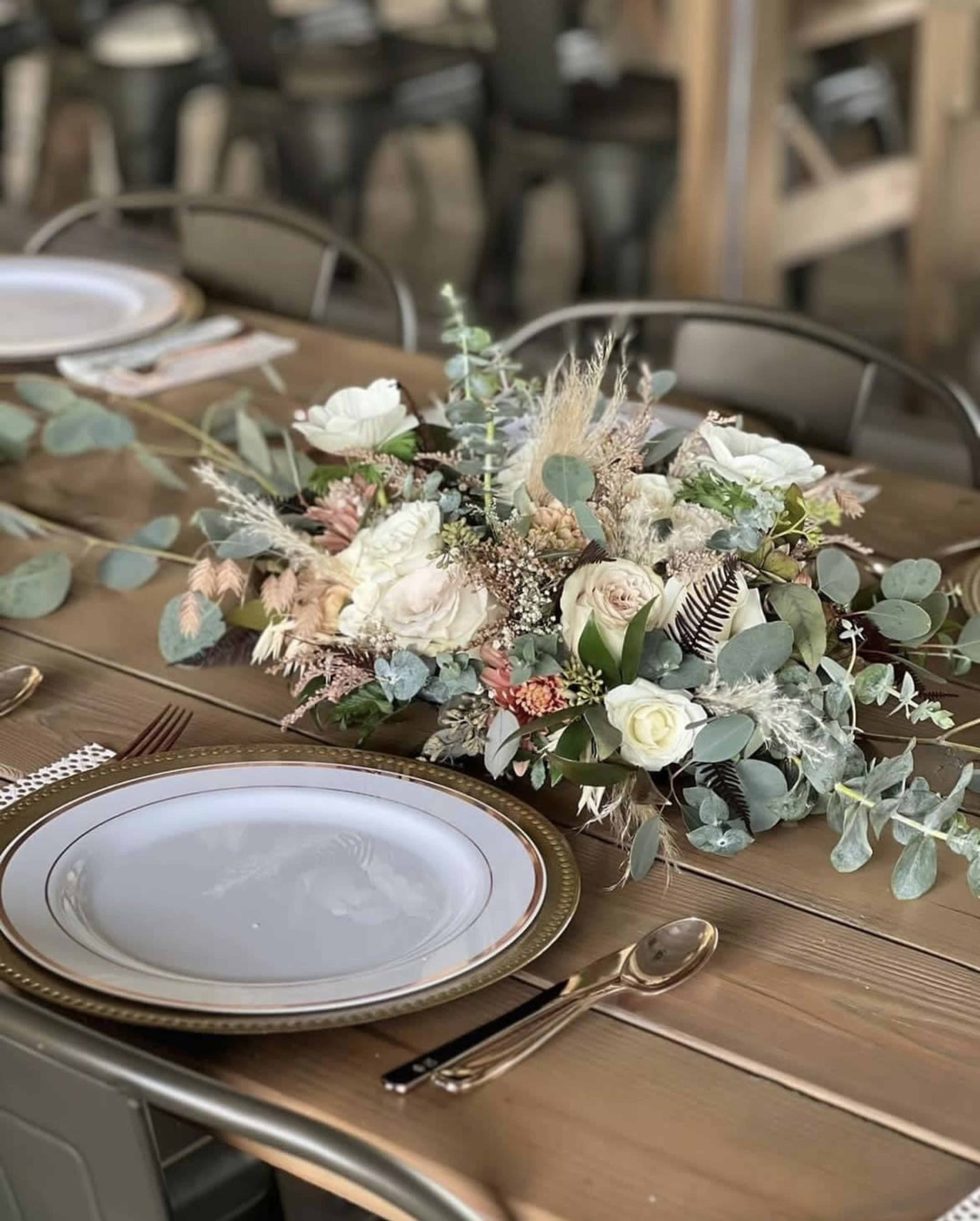 A floral centerpiece made of various dried and fresh flowers is placed on a wooden dining table set with white plates and shiny cutlery.