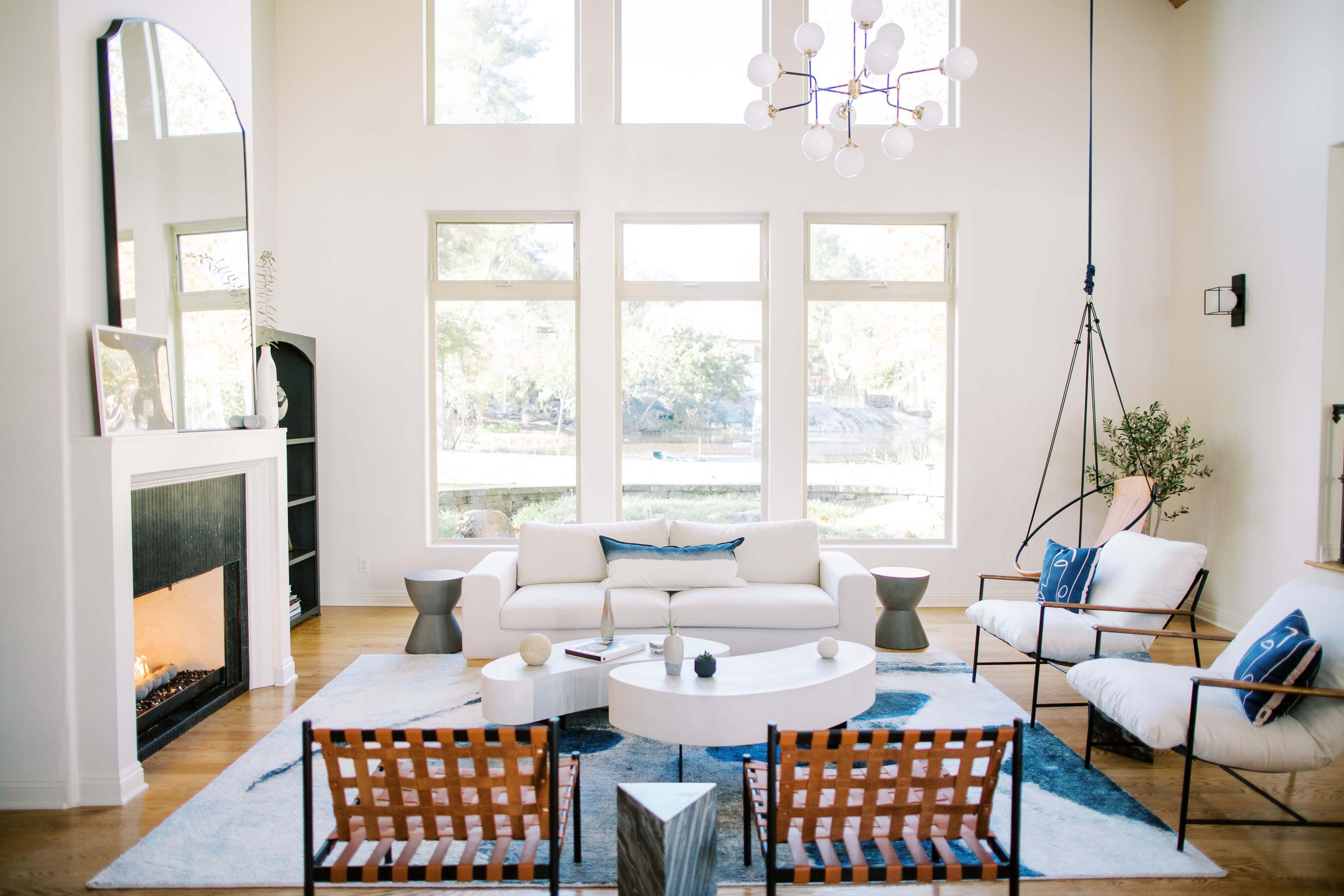 A bright living room features large windows, a white sofa, a round coffee table, and a cozy fireplace.
