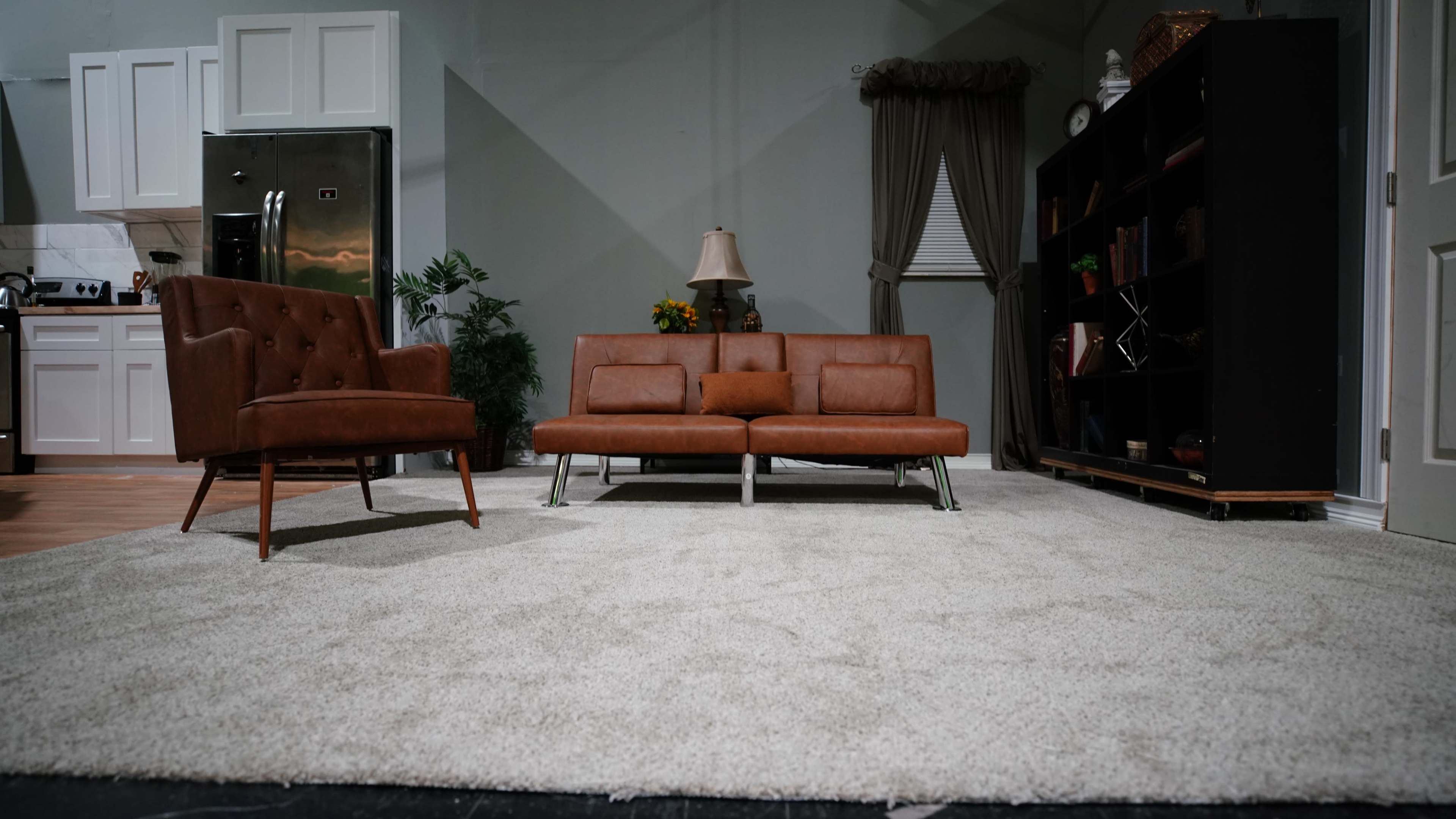 A living room set features a brown leather sofa and armchair, a lamp, and a black bookshelf, all arranged on a light-colored carpet.