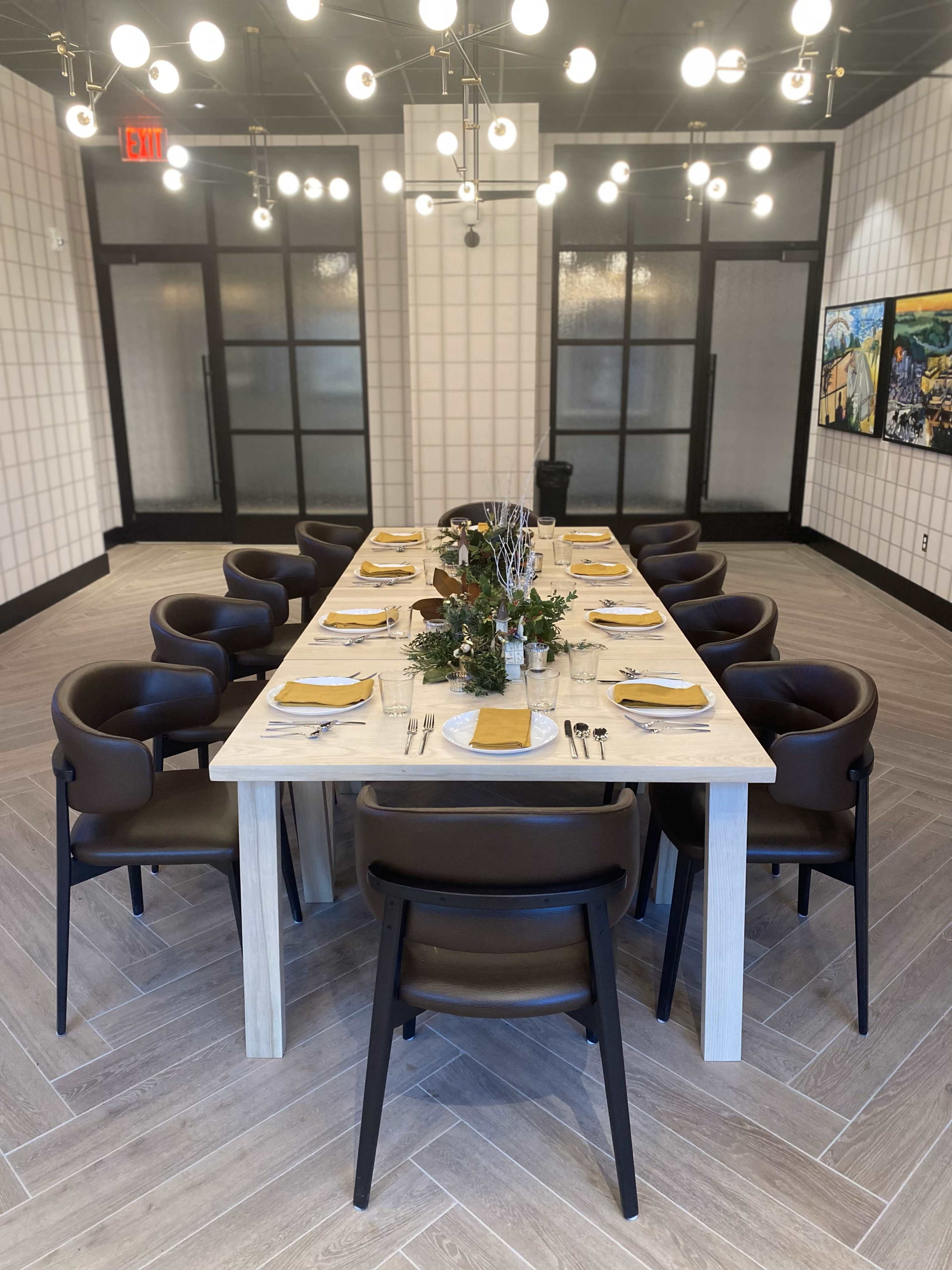 A long, rectangular dining table is set with dishes and utensils, surrounded by dark chairs in a well-lit modern room.