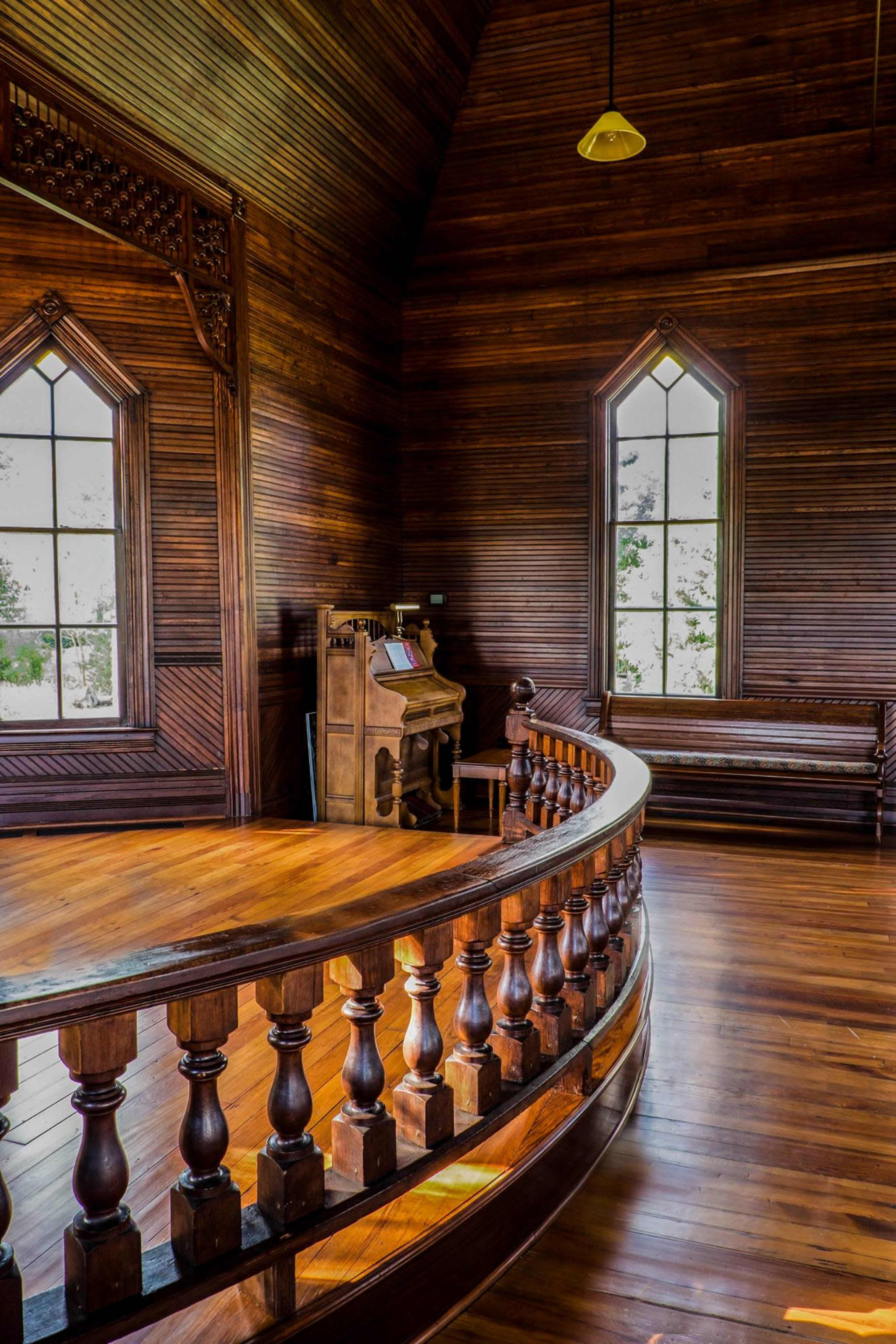 The image shows a wooden interior of a room featuring large windows, a curved railing, and an antique organ placed against the wall.