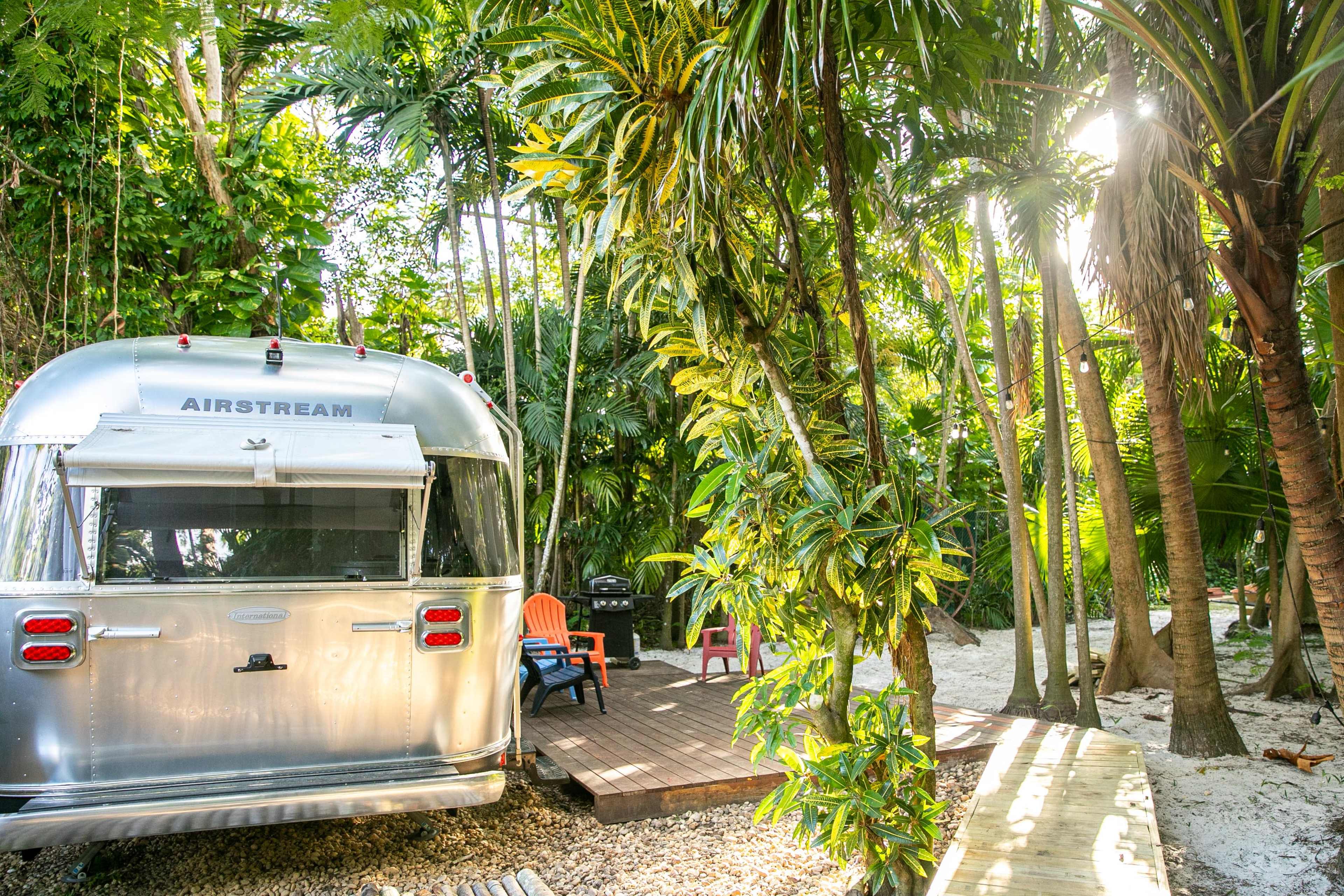 A silver Airstream trailer is parked among lush tropical trees, with a small wooden deck and chairs nearby.