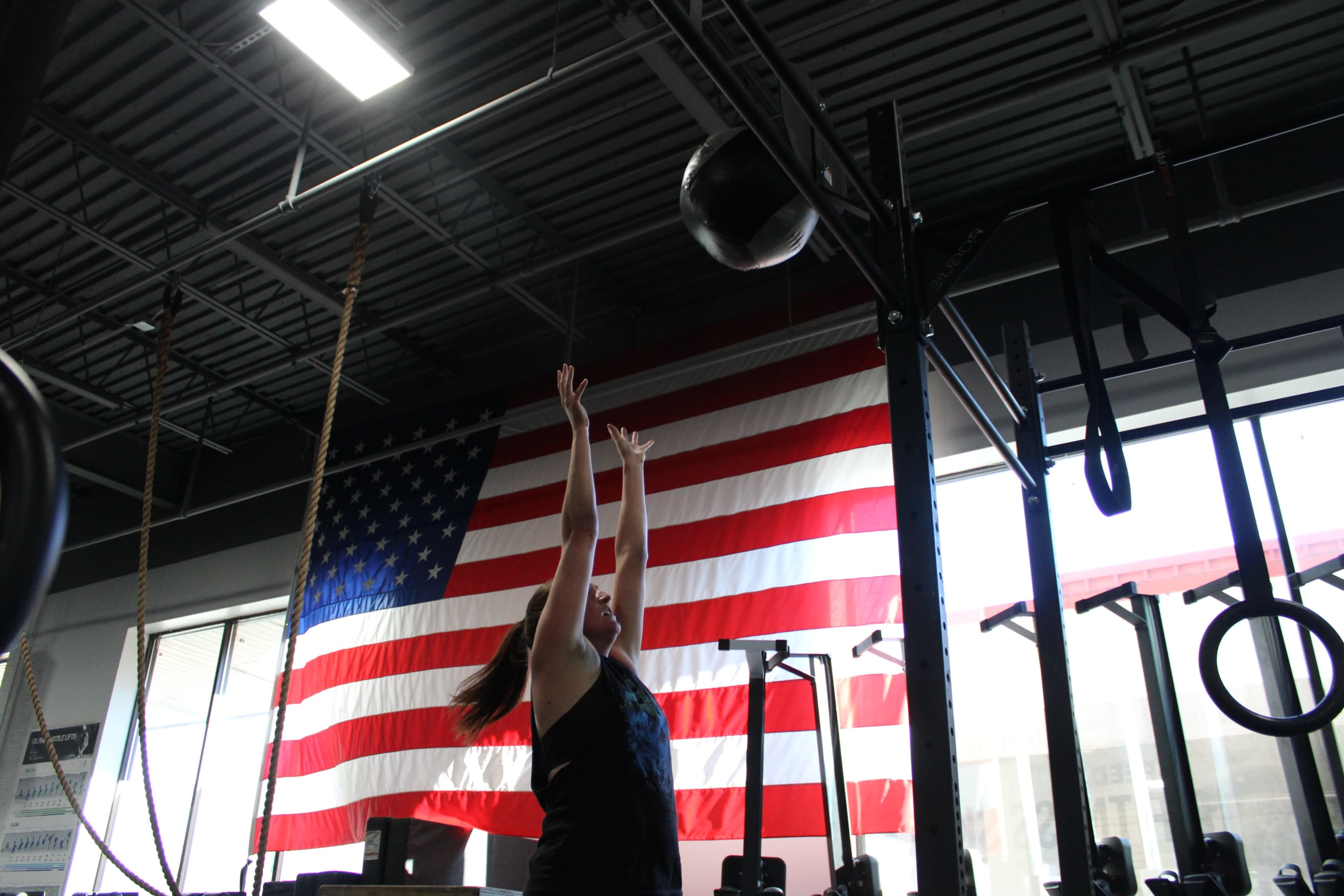 A person in a gym tosses a medicine ball upward in front of a large American flag.
