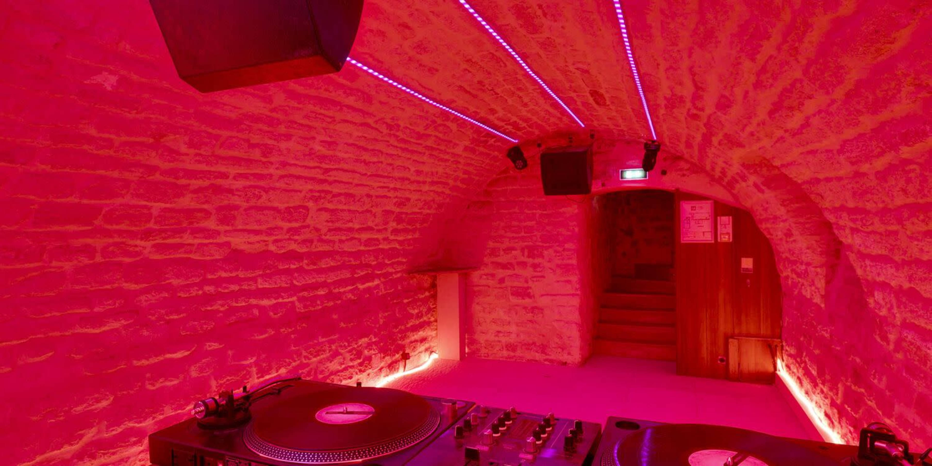 A dimly lit, arched room with stone walls, featuring a DJ setup and pink LED lighting along the ceiling.