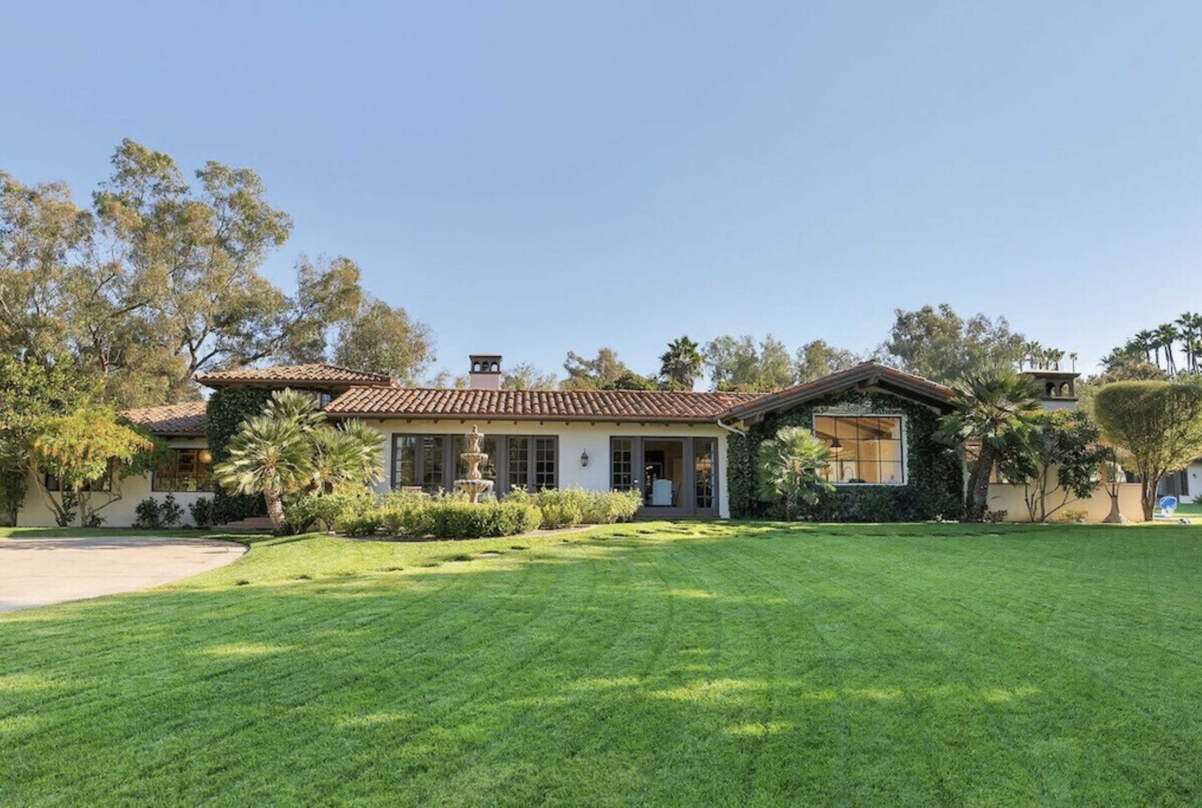 A single-story house with a tiled roof is situated on a well-maintained lawn surrounded by trees and shrubs.