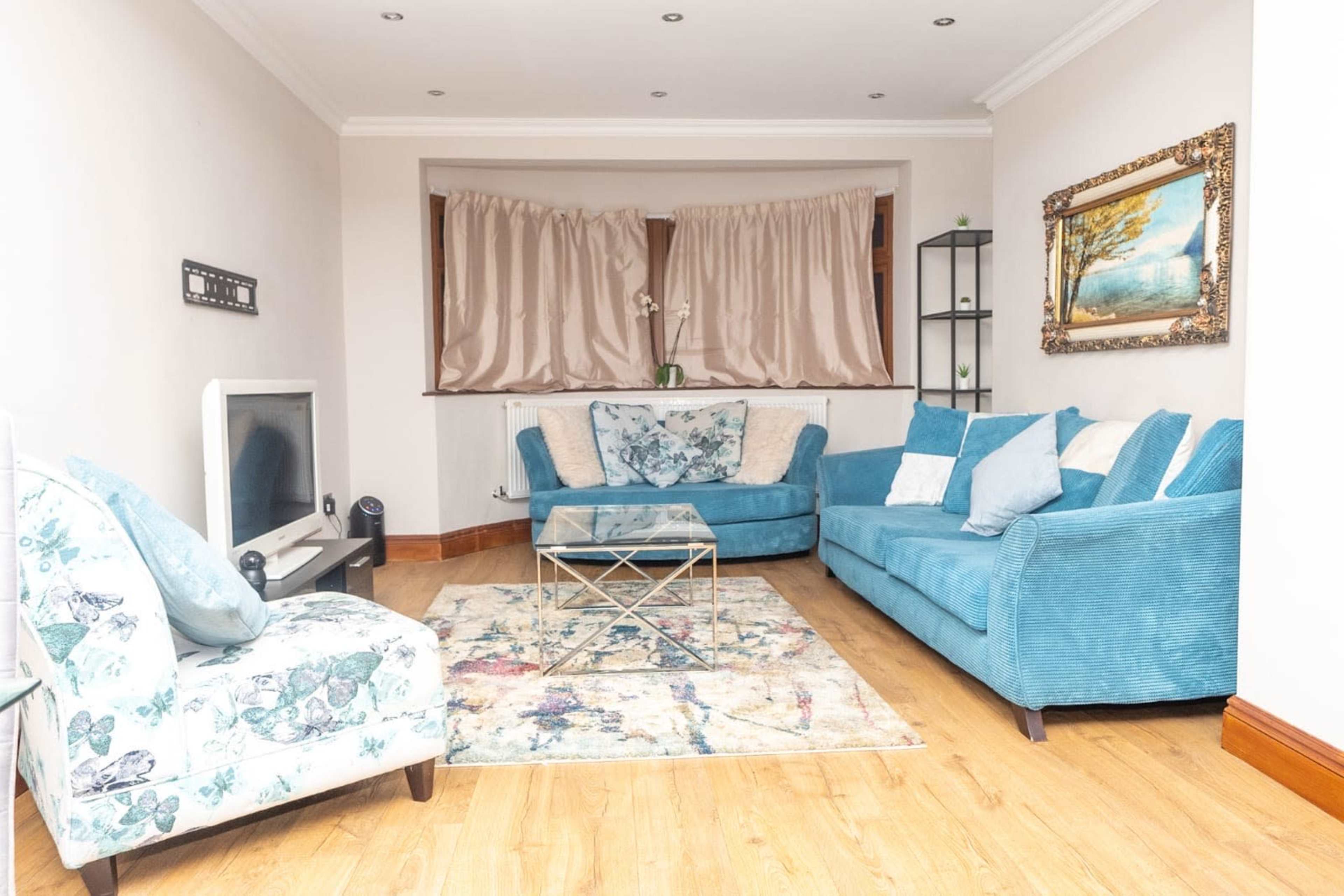 The living room features a blue sofa set, a glass coffee table, and a floral armchair, arranged around a colorful area rug.