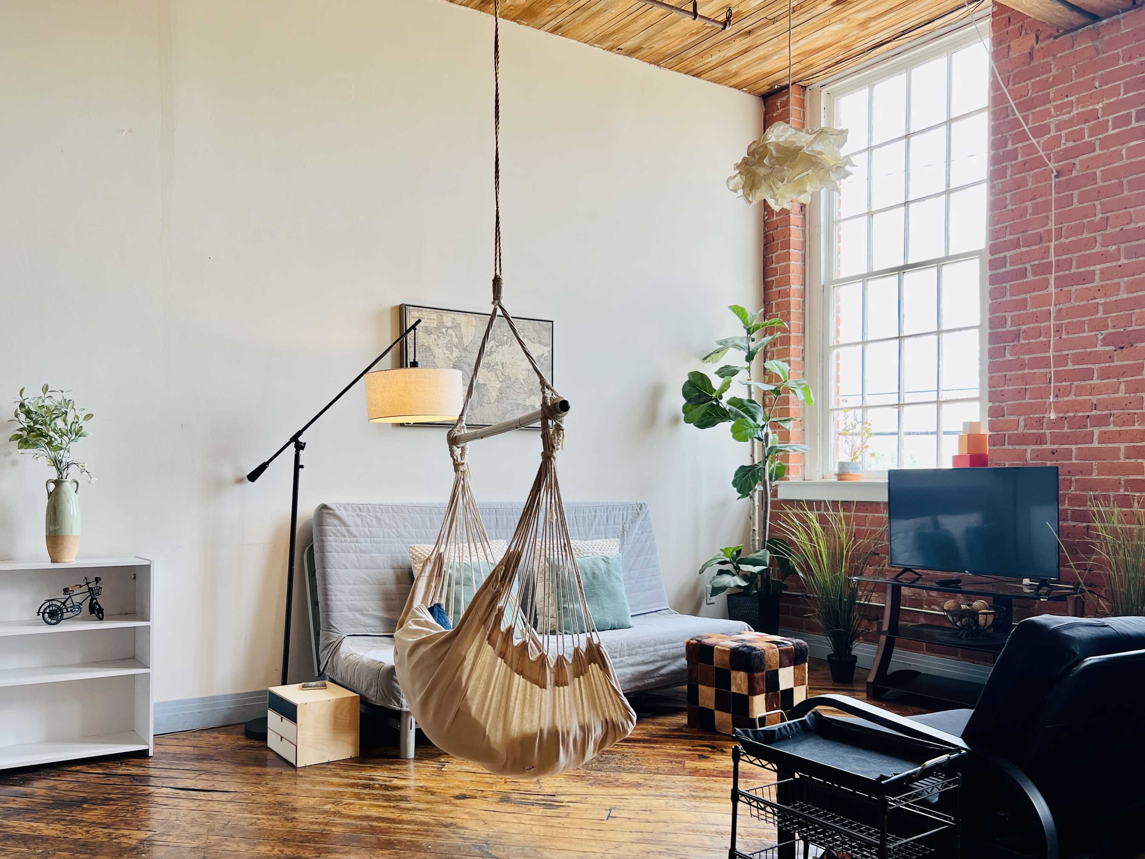 The image shows a cozy living space featuring a hammock chair, a gray sofa bed, a small television on a stand, and plants against exposed brick walls.