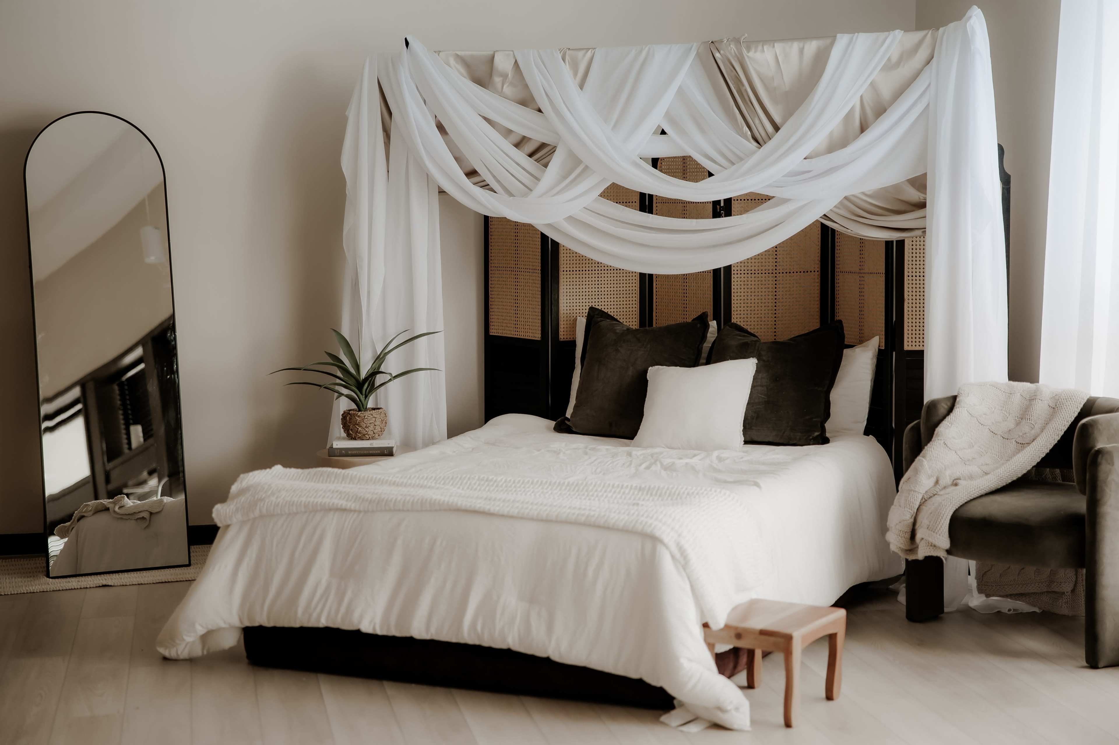 The image depicts a neatly arranged bedroom featuring a large bed with draped white fabric above, flanked by a mirror and a potted plant.