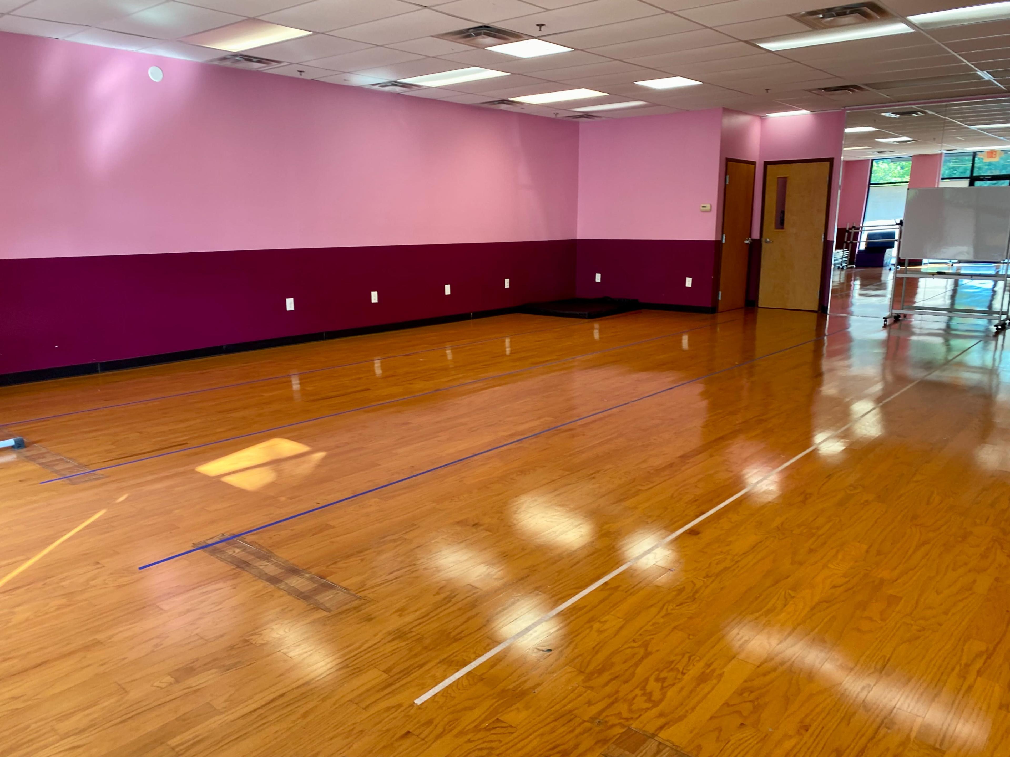 A spacious room with polished wooden floors, painted pink and maroon walls, and outlined flooring markers for activities.