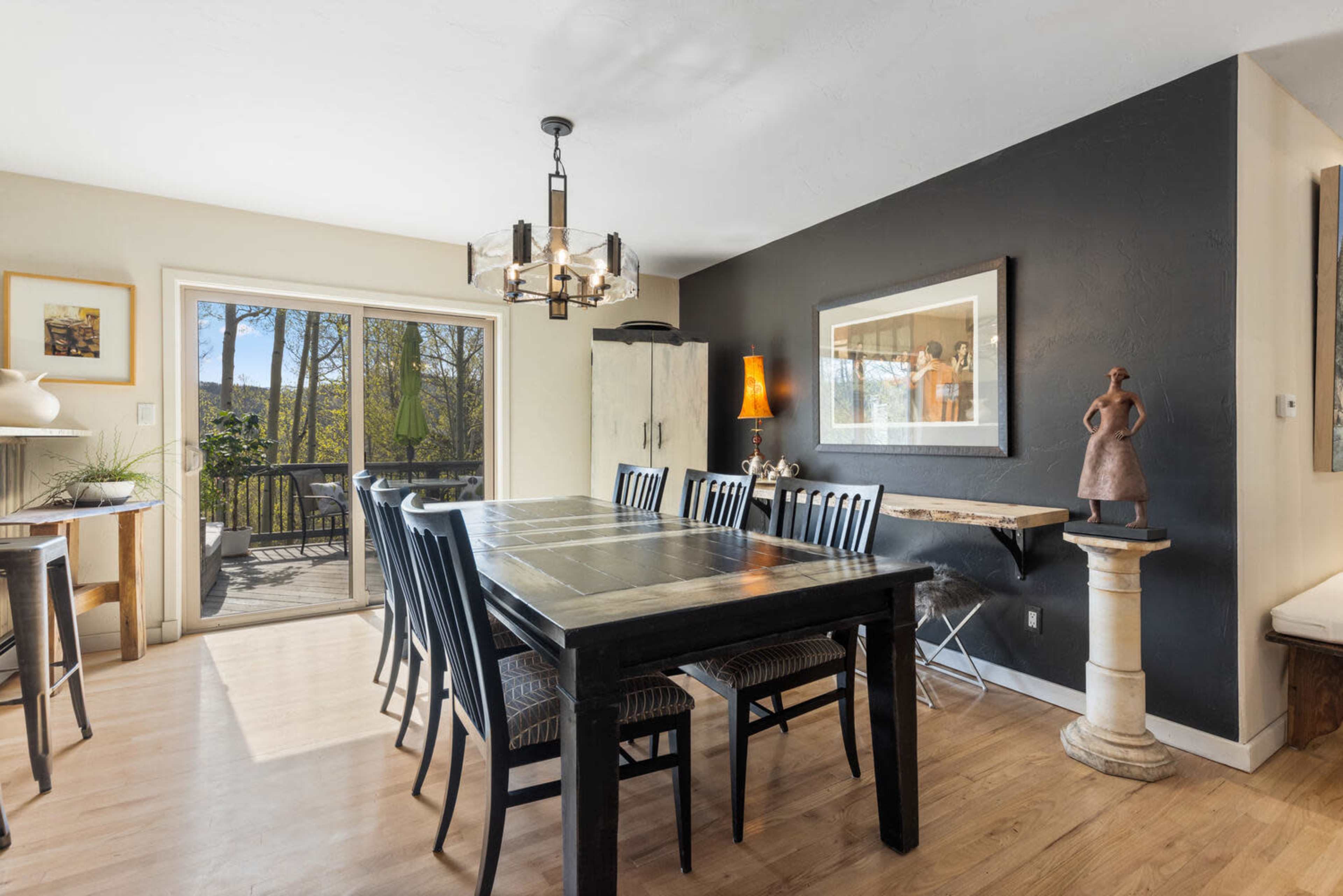 A dining area features a large black table surrounded by black chairs, with a sliding glass door leading to a deck, and art displayed on the walls.
