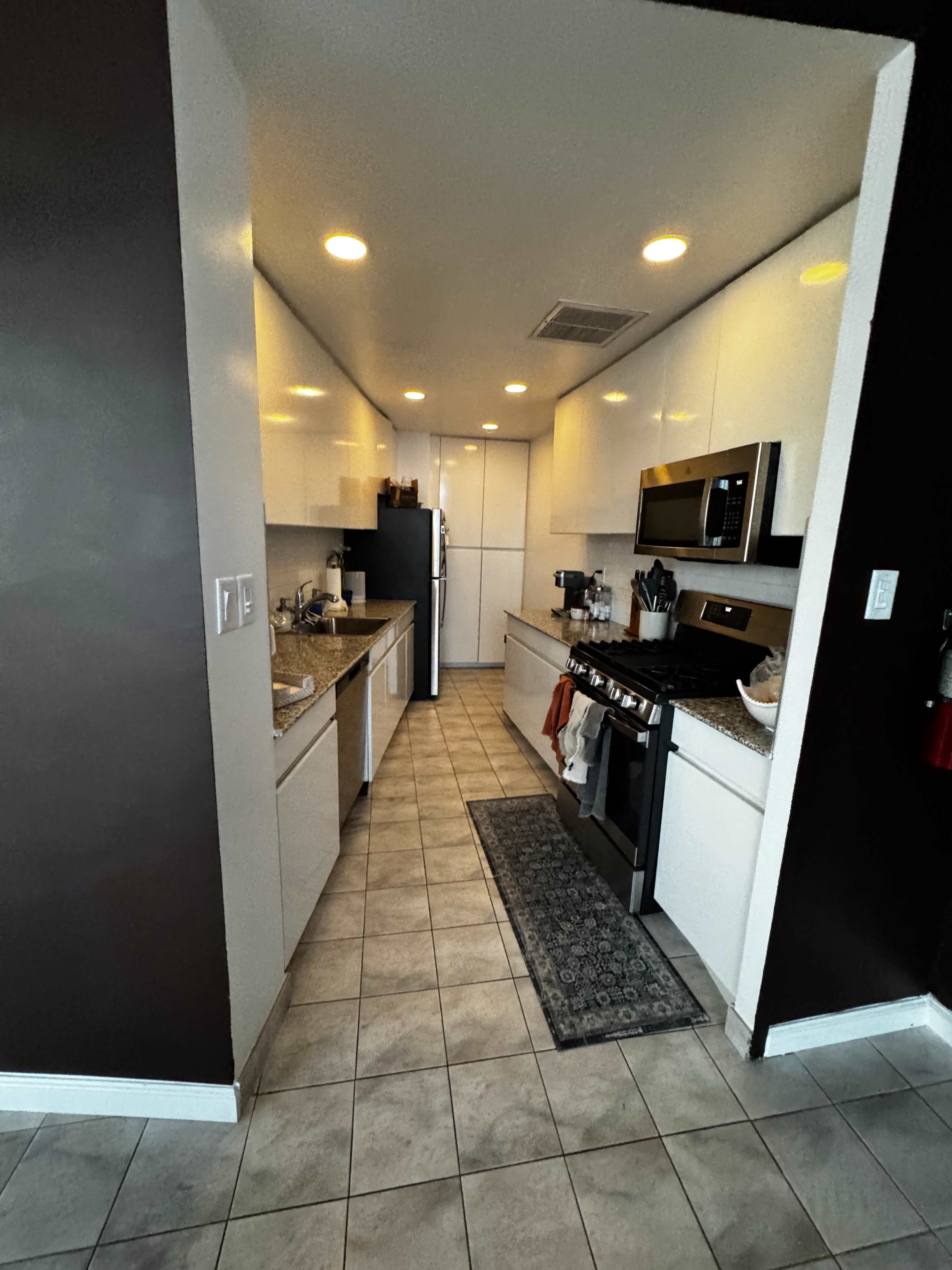 A modern kitchen features sleek white cabinetry, stainless steel appliances, and a tile floor with a runner mat along the center.