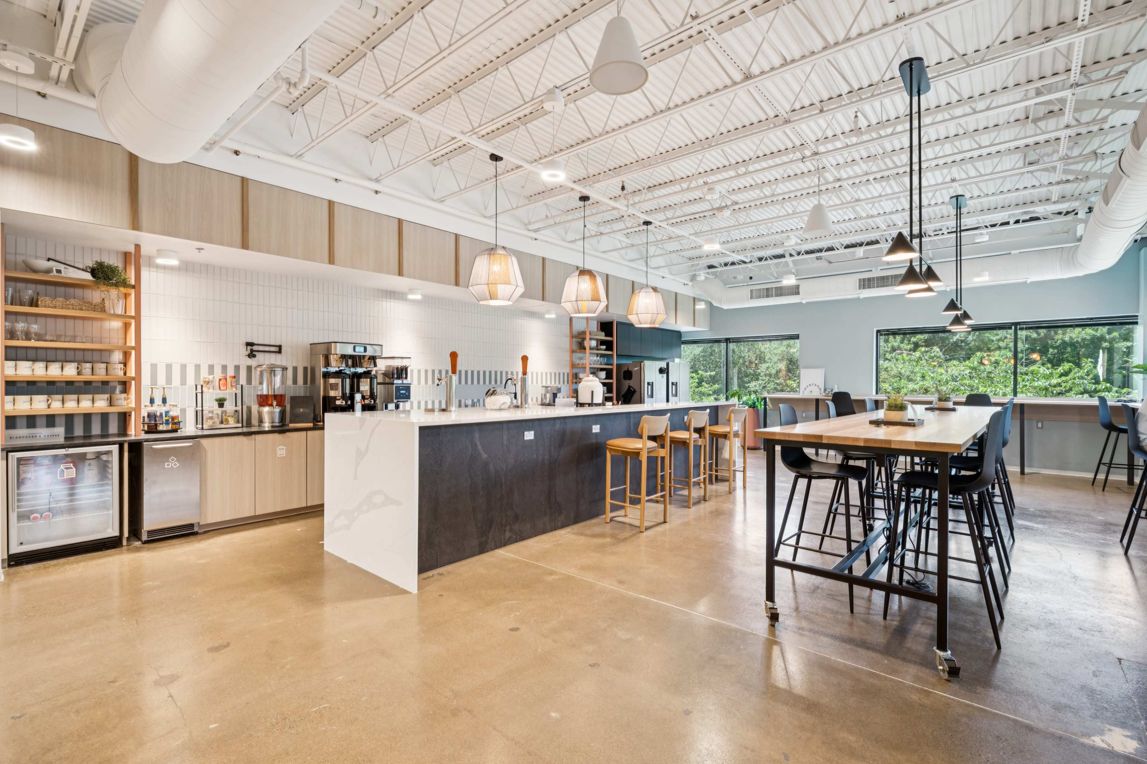 The image shows a modern kitchen area with a long countertop, seating, and various appliances, illuminated by pendant lights and natural light from large windows.