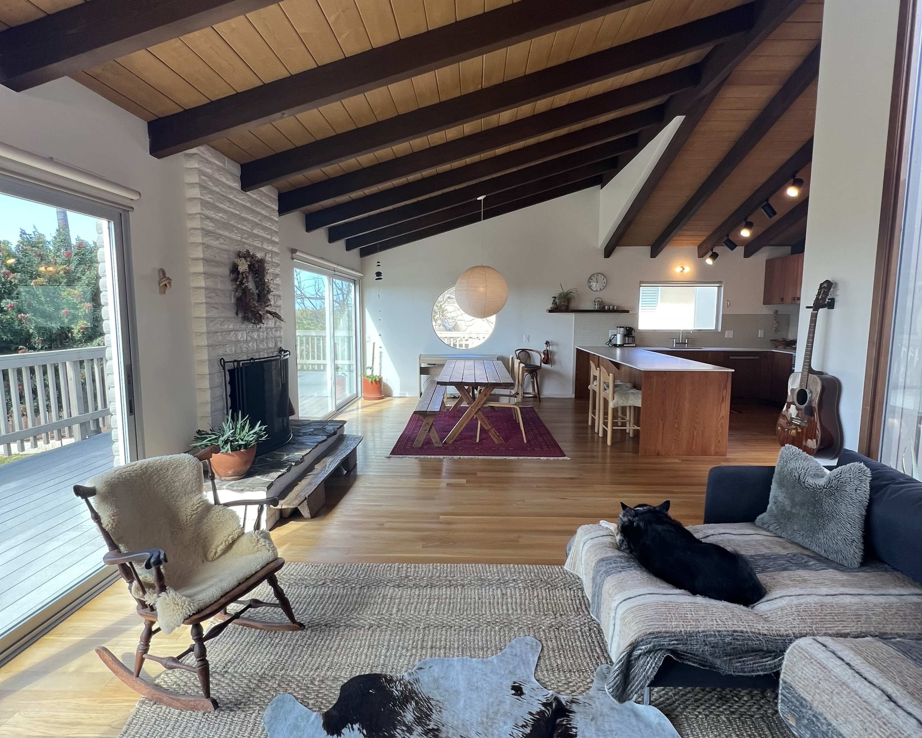 The image shows a spacious living area featuring a seating arrangement, dining area, and an open kitchen with wooden beams and large windows.