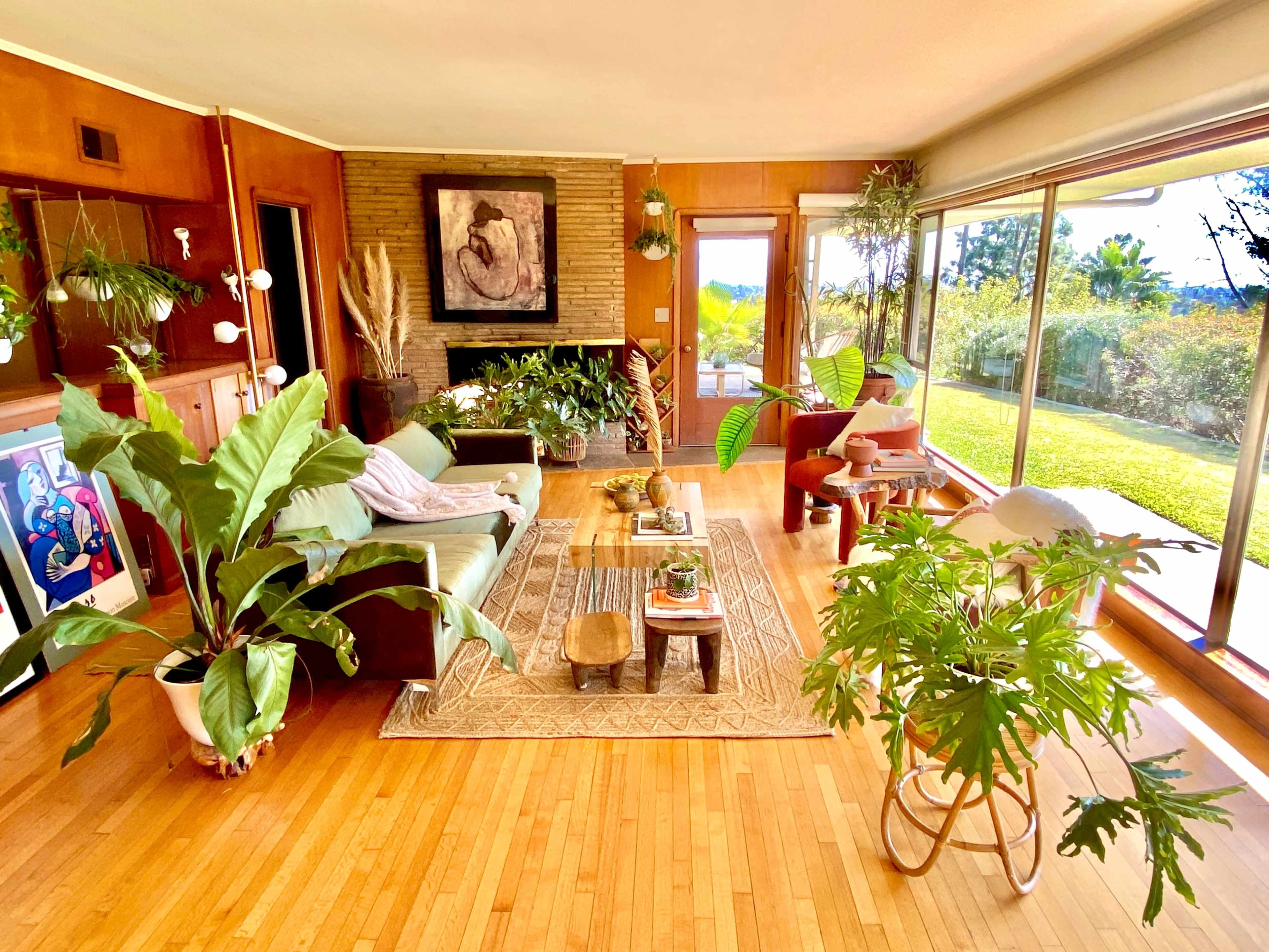 The image shows a mid-century modern living room with large windows, hardwood floors, various indoor plants, and a cozy seating area.