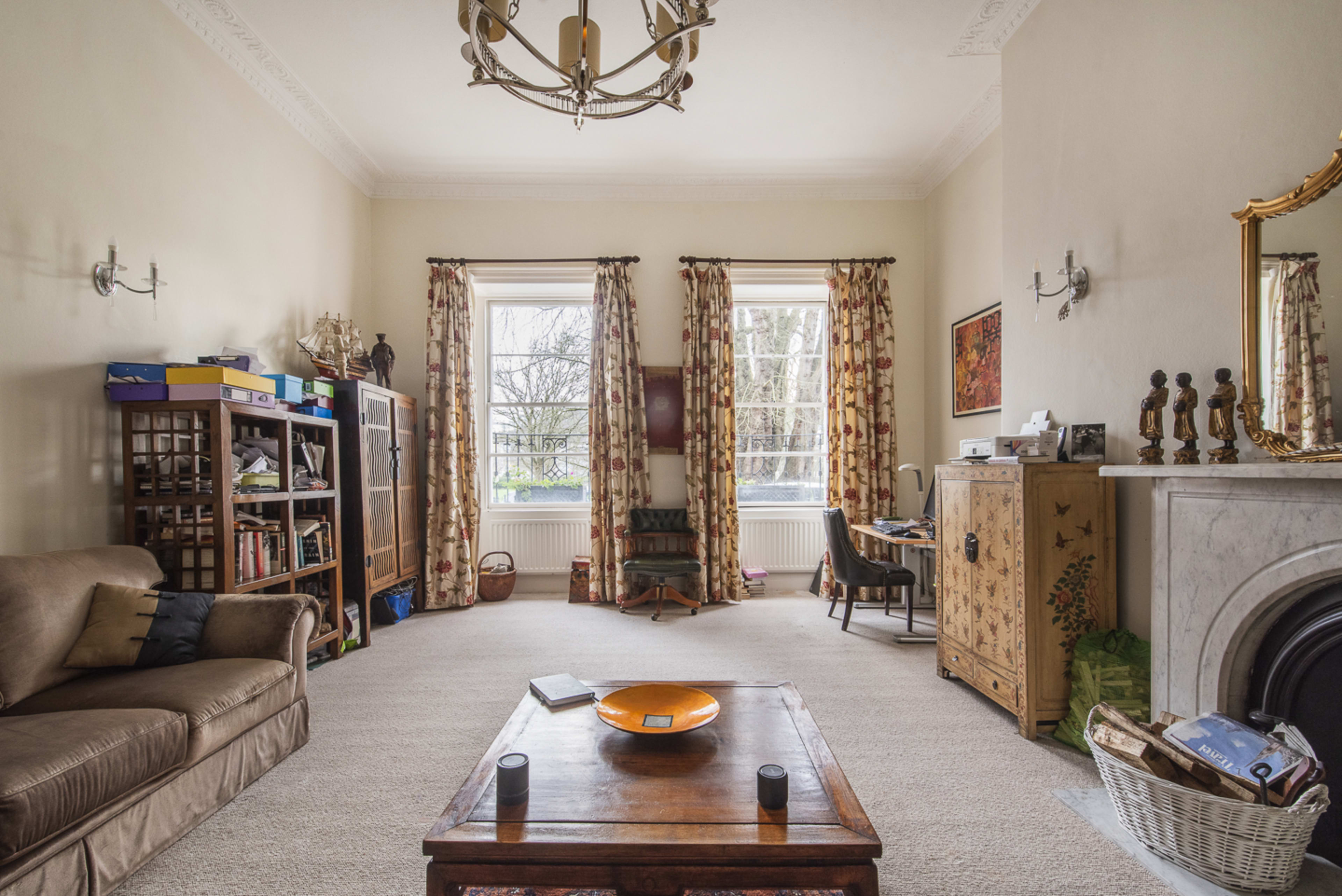 The image depicts a spacious living room with a brown sofa, a wooden coffee table, bookshelves, and large windows adorned with patterned curtains.